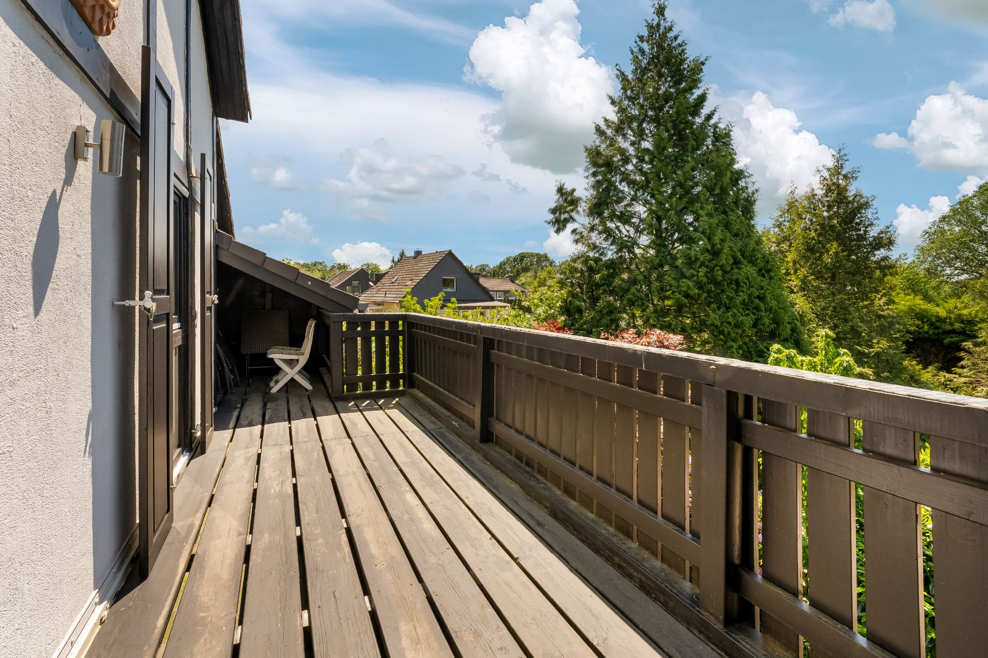 Holzbalkon mit Stuhl, angrenzend an ein Wohnhaus, umgeben von Bäumen und Gärten.