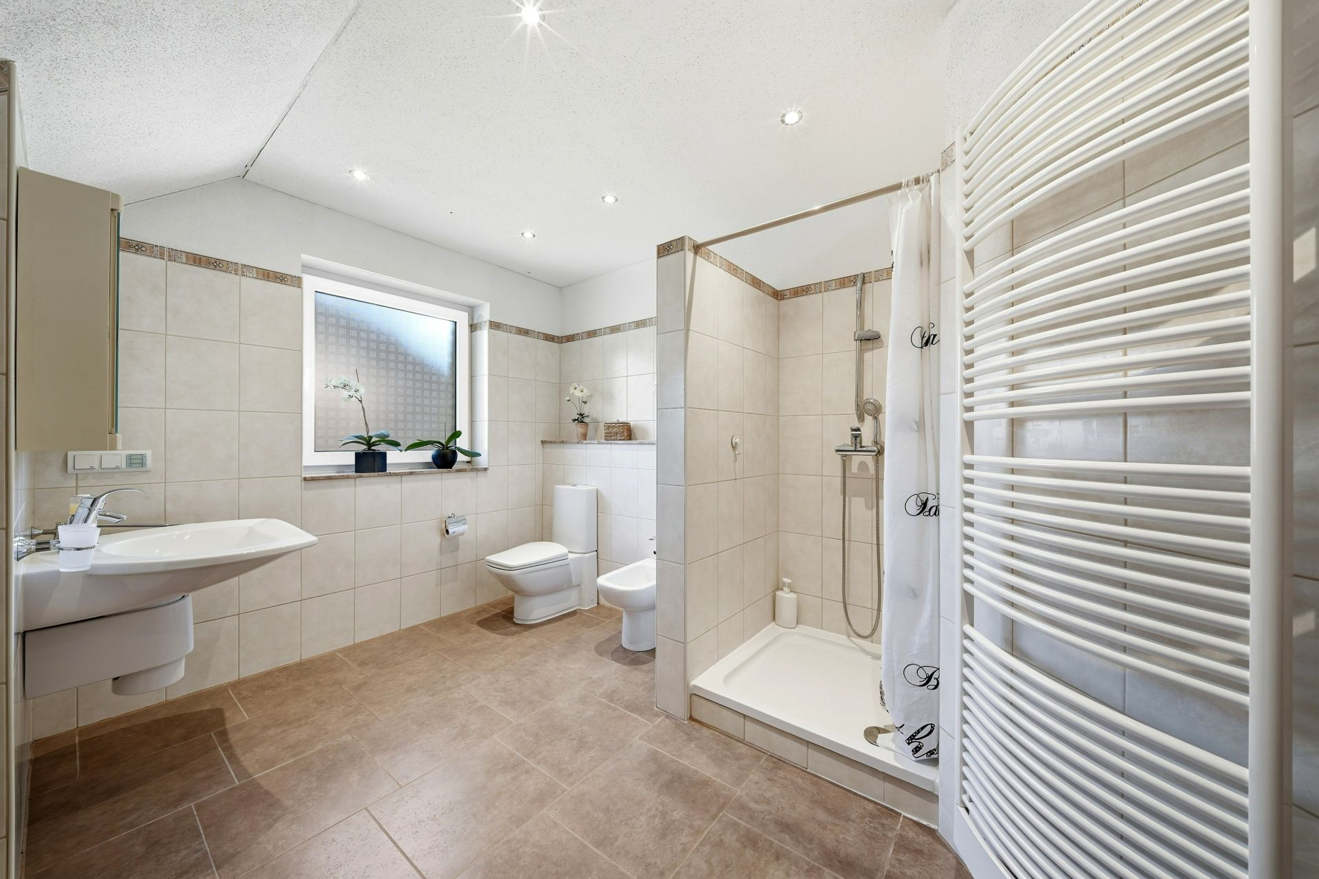Modernes Badezimmer mit Dusche, Waschbecken, WC, Bidet, Fenster und Handtuchheizkörper.