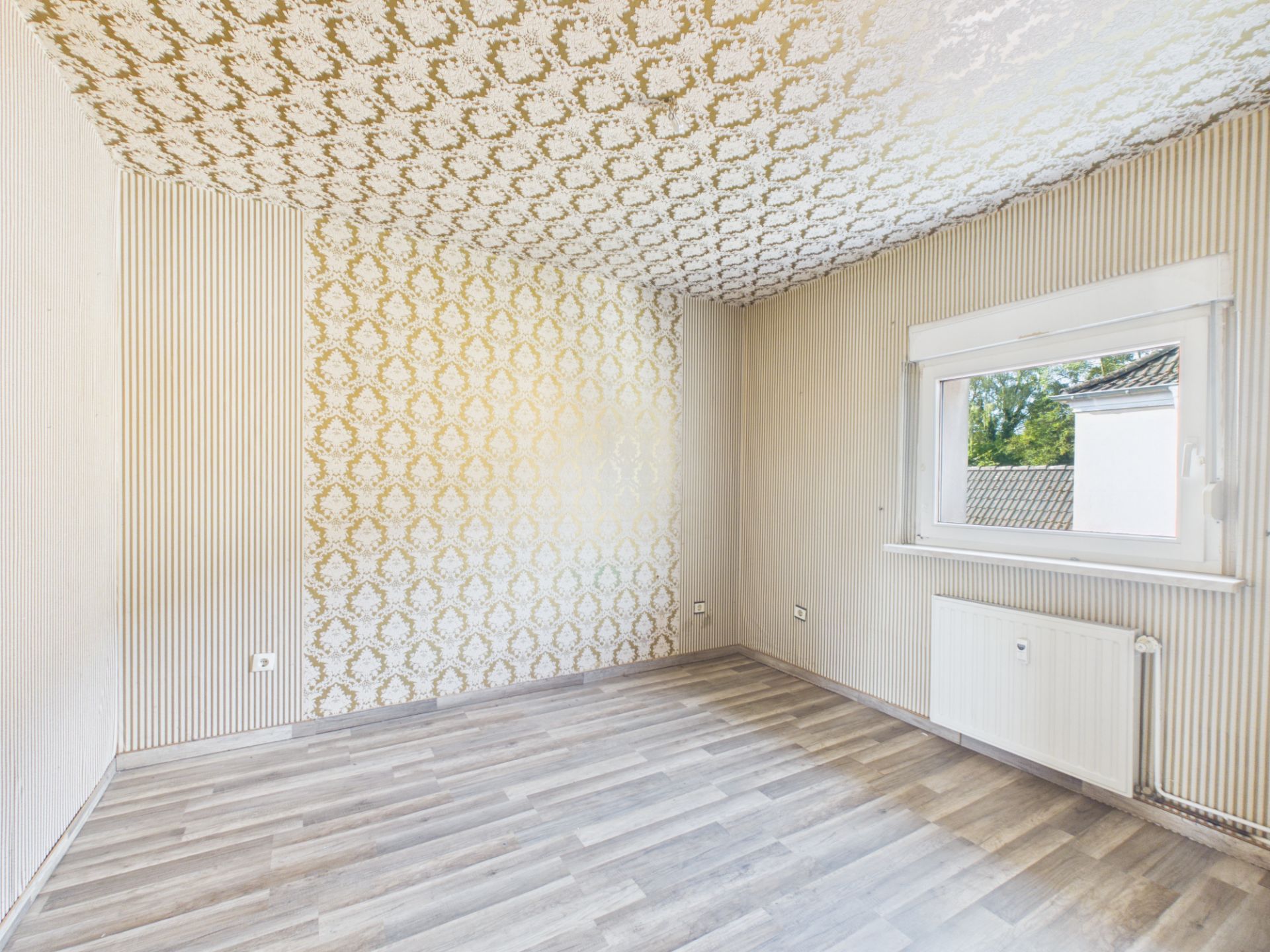 Leeres Zimmer mit gemusterten Tapeten, Fenster, Heizkörper und hellem Boden.