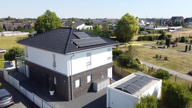 Modernes Wohnhaus mit dunklem Dach und Solarpanels, Garage mit Solarpanels, umgeben von Grünflächen.