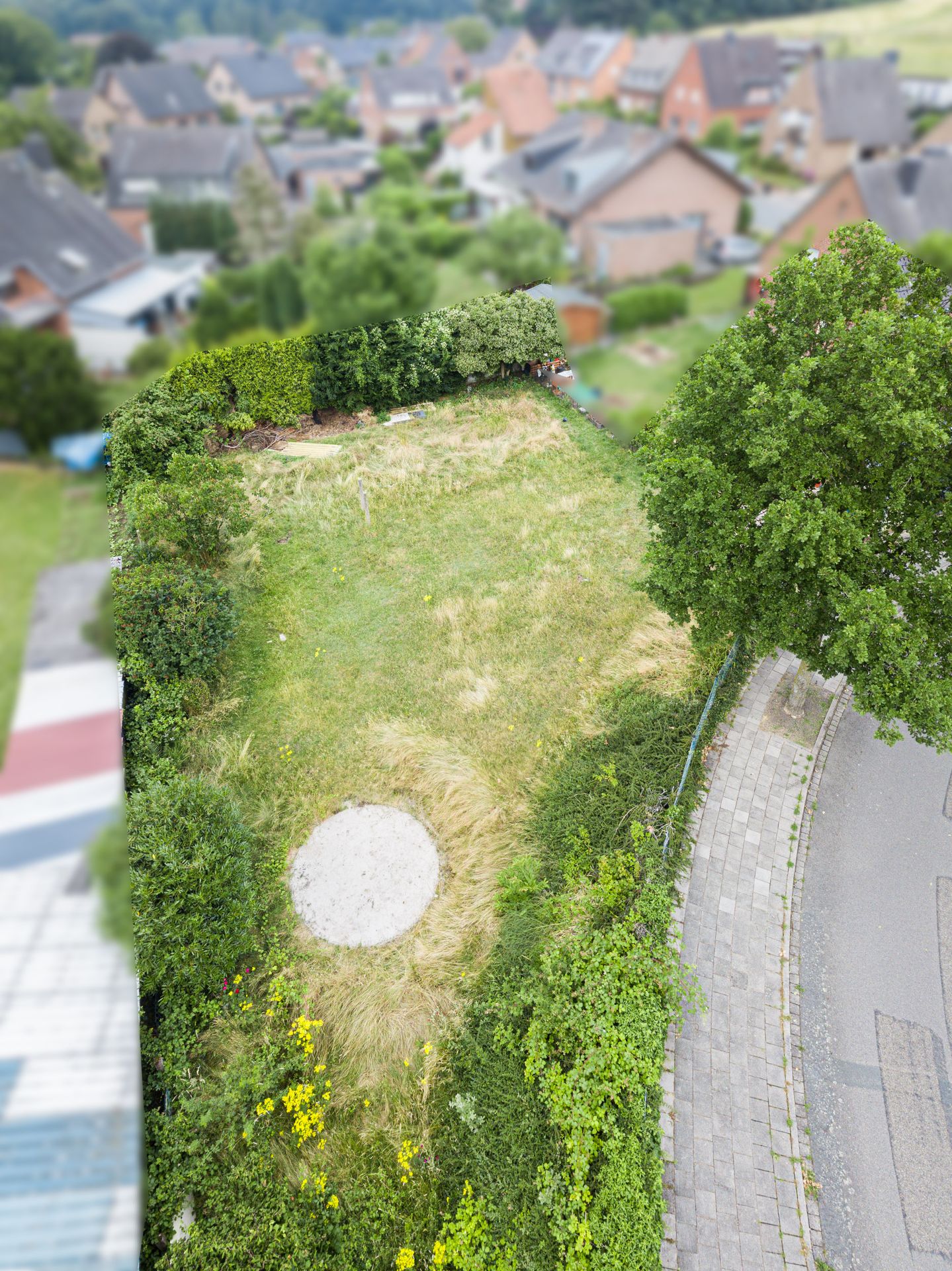 Luftaufnahme eines umzäunten Gartens mit Rasen und rundem Sandplatz, daneben eine gepflasterte Straße.