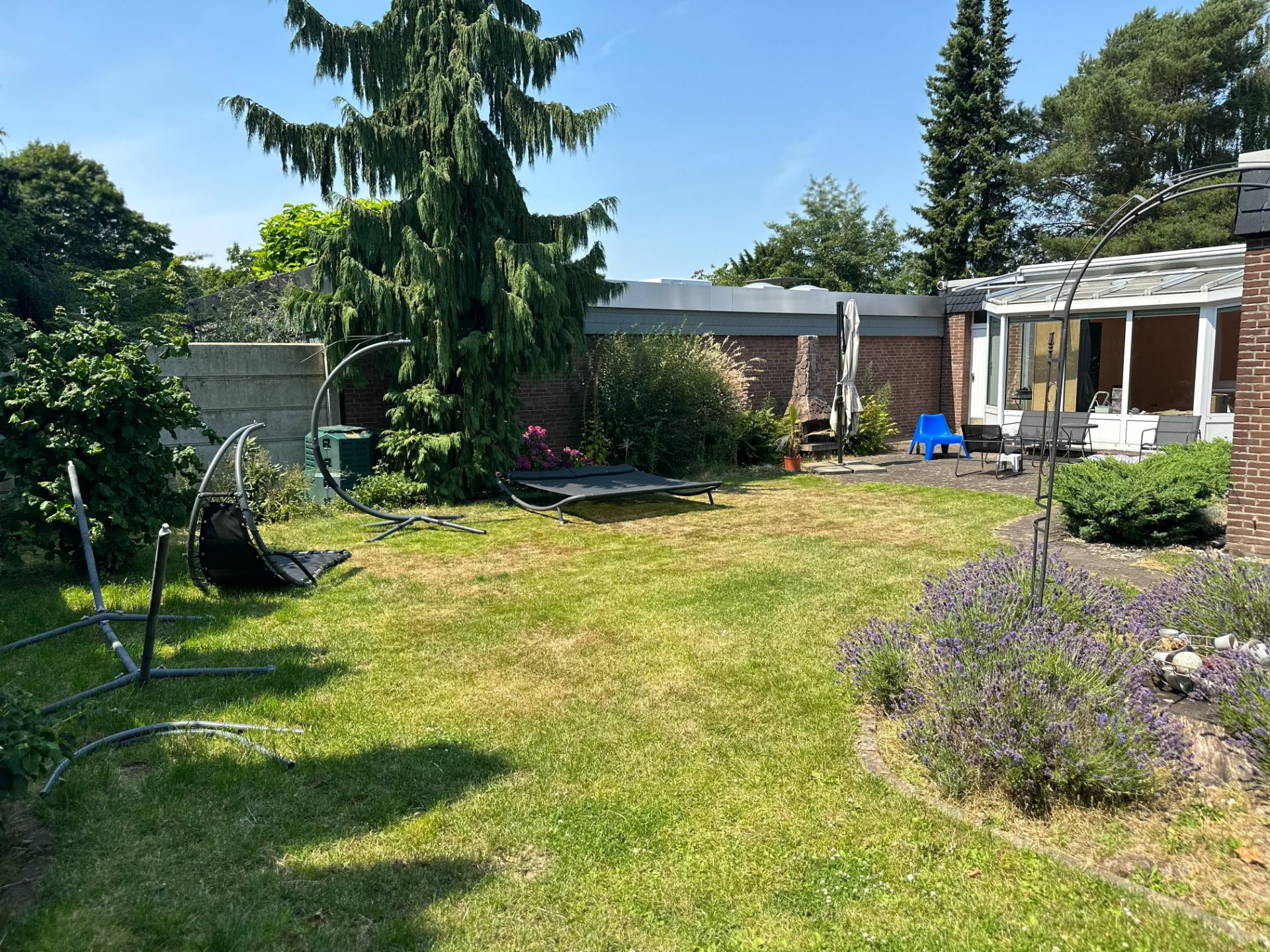 Garten mit Rasen, Bäumen, Sträuchern und Terrasse an einem Wohnhaus bei Sonnenschein.