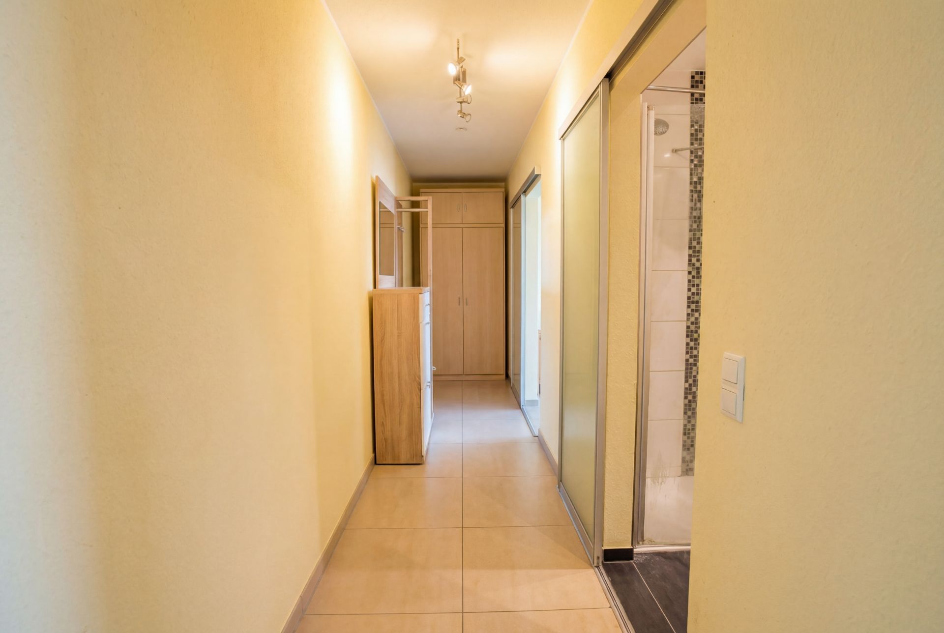 Heller Flur mit Fliesenboden, Spiegel und Holzschrank, rechts Zugang zu einem Badezimmer.