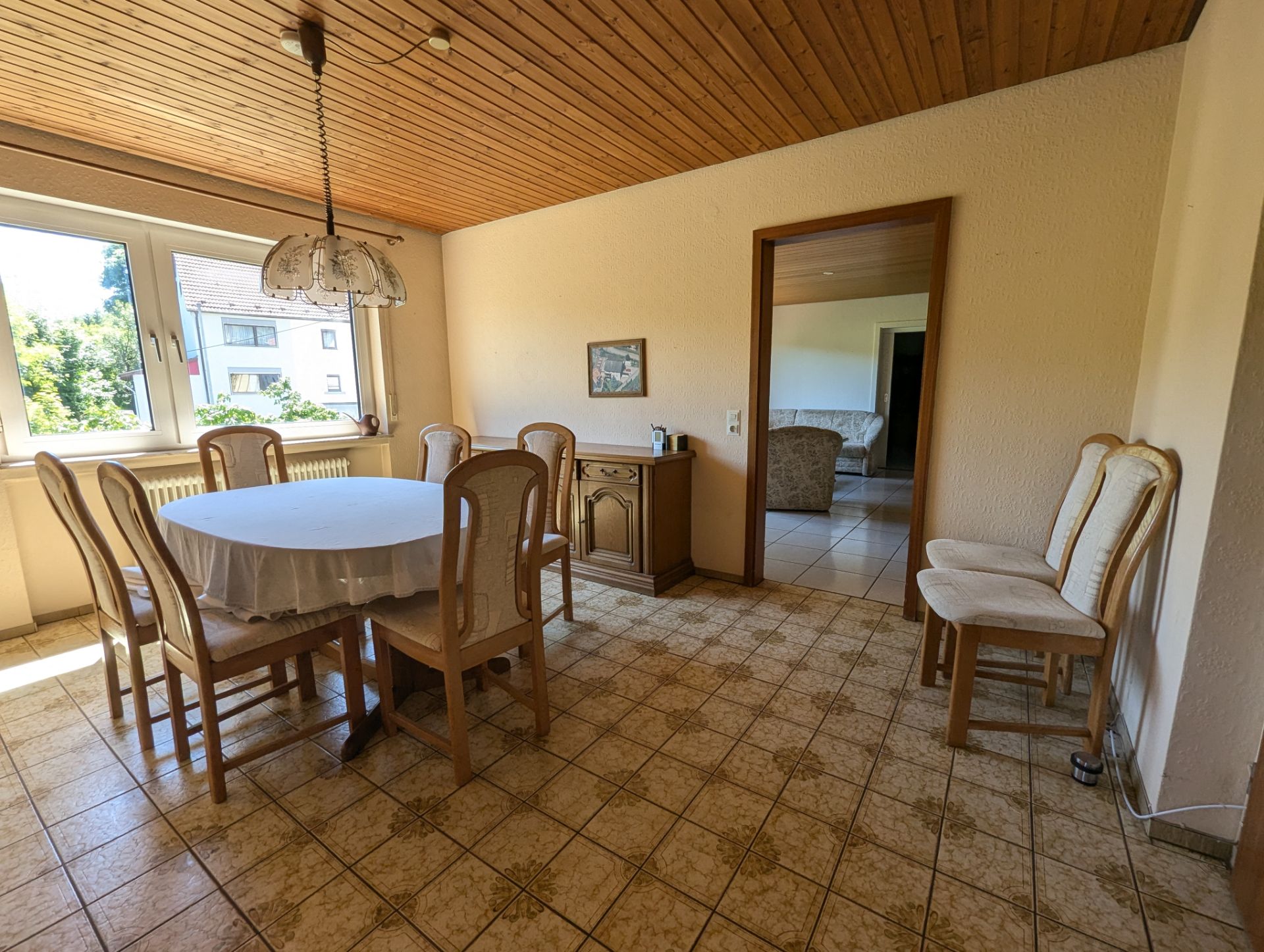 Esszimmer mit rundem Tisch, sechs Stühlen, Kommode und großem Fenster, Blick ins Wohnzimmer.