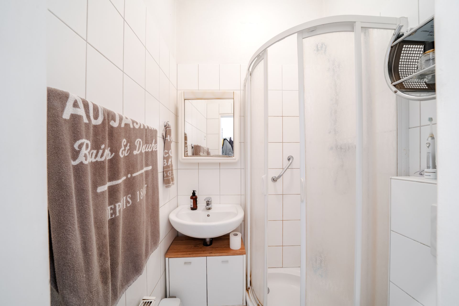 Kleines Badezimmer mit Dusche, Waschbecken, Spiegel und Handtuch.