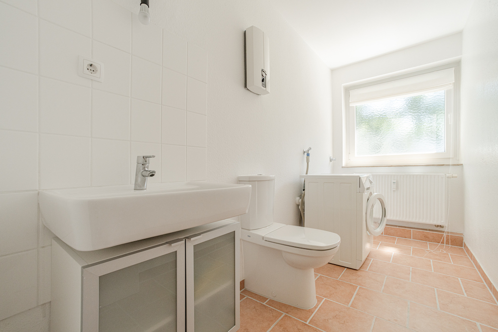 Helles Badezimmer mit Waschbecken, Toilette, Waschmaschine und Fenster mit Rollo.