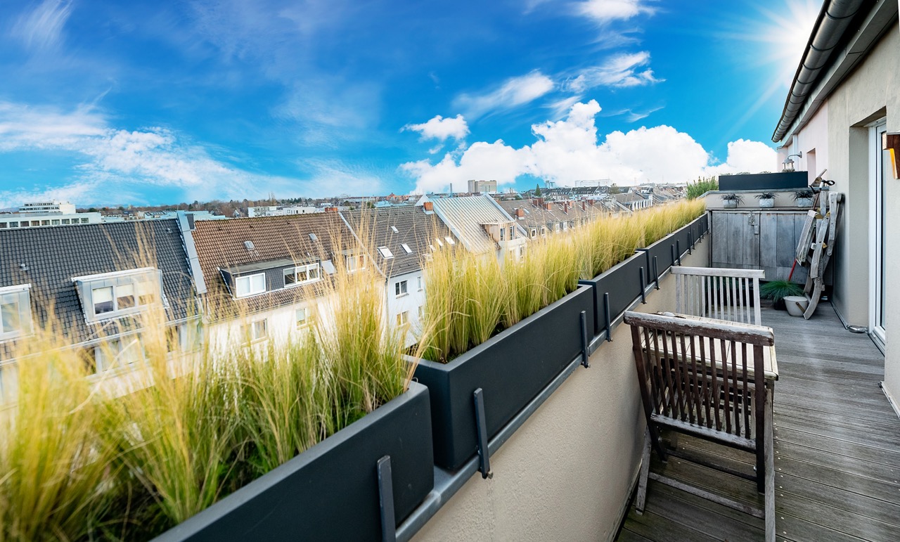 Langer Balkon mit Holzmöbeln, Pflanzkästen und Blick auf Dächer in einer Wohngegend.