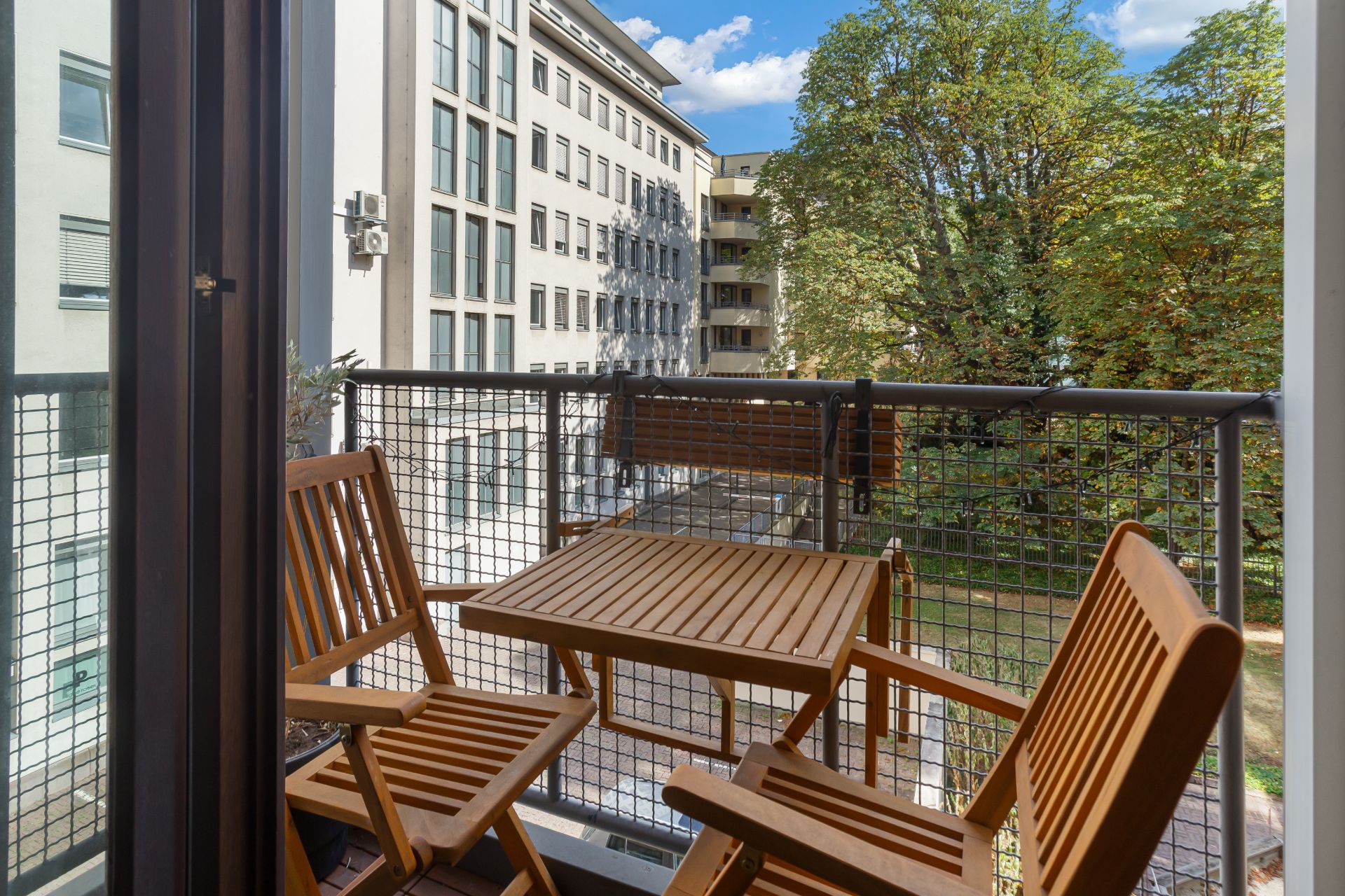 Kleiner Balkon mit Holztisch und drei Holzstühlen, Blick auf Bäume und Wohngebäude.