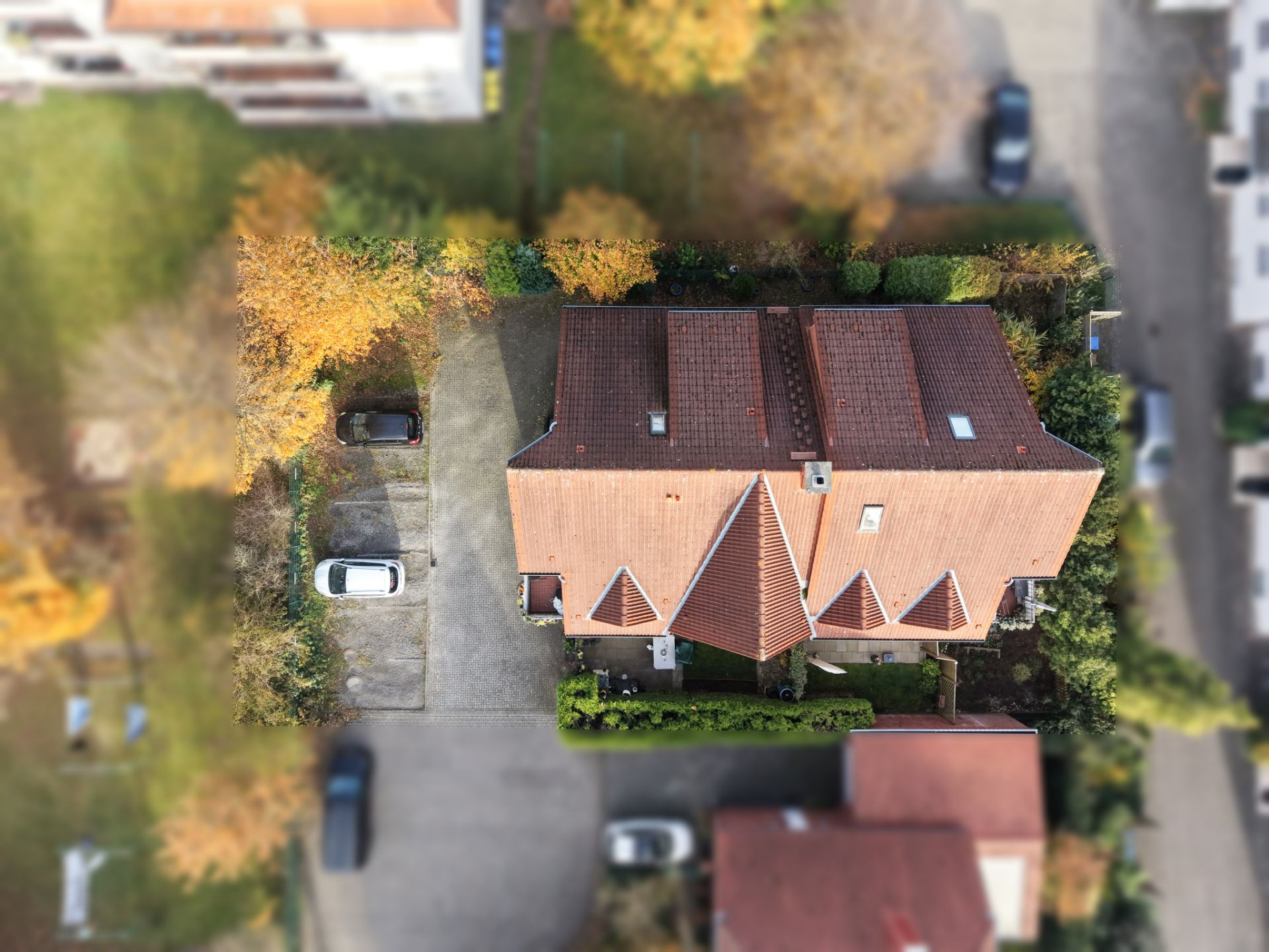 Luftaufnahme eines Einfamilienhauses mit rotem Dach und angrenzender Einfahrt mit geparkten Autos.