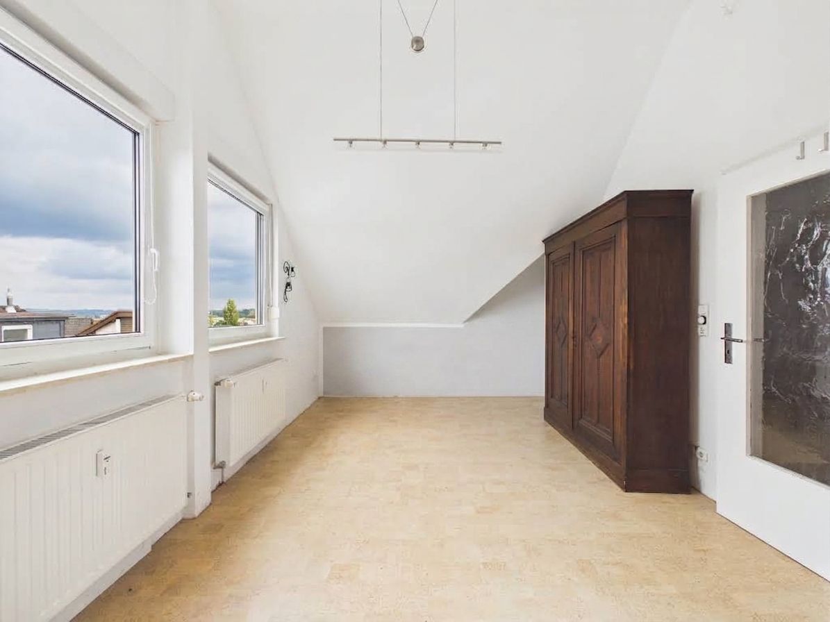Helles Zimmer mit zwei Fenstern, Dachschräge, Holzboden und großem dunklen Schrank.