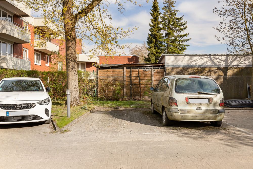 Zwei geparkte Autos auf einem Parkplatz vor Wohngebäuden, Bäume und Zaun im Hintergrund.