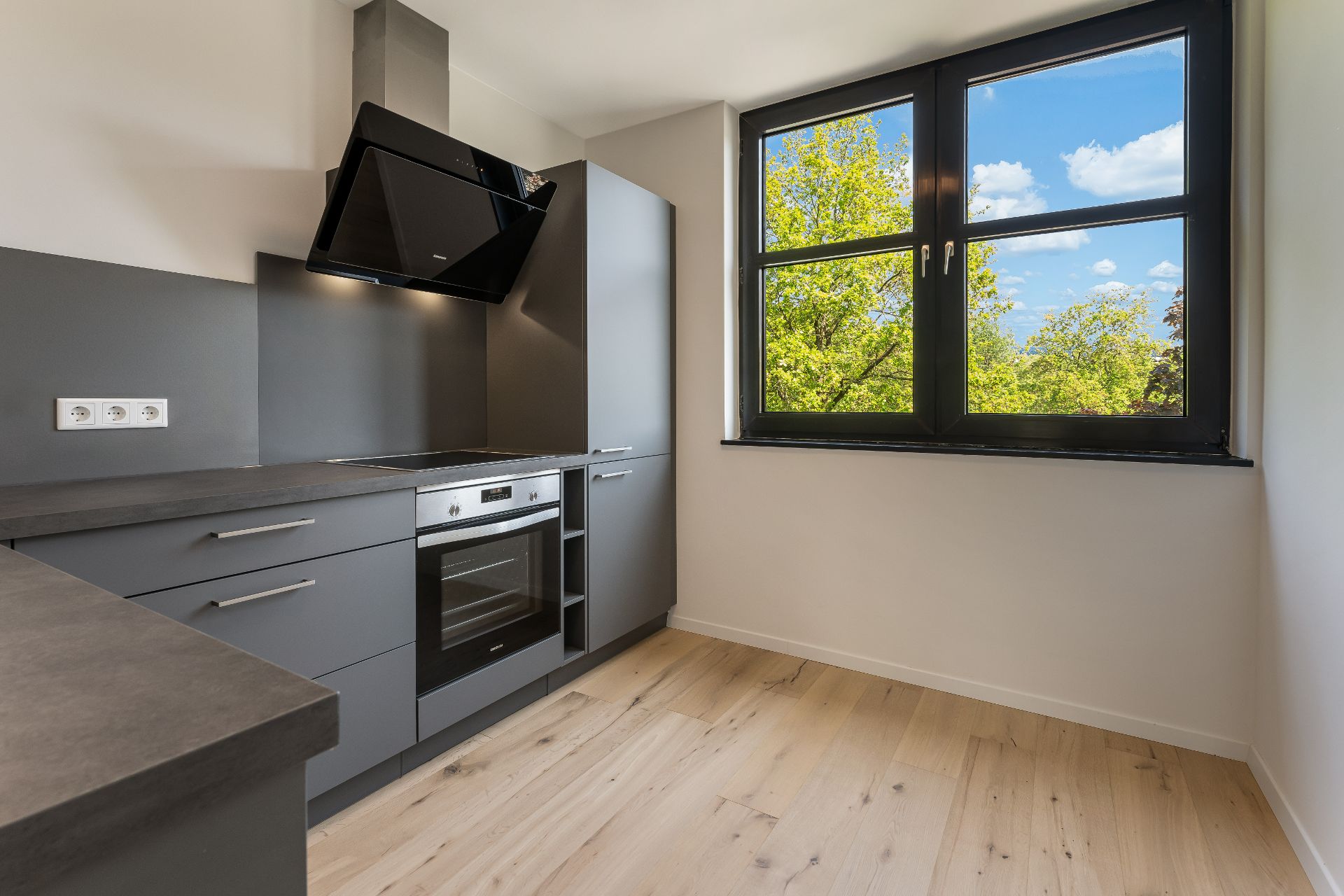 Moderne Küche mit grauen Schränken, Herd, Backofen und großem Fenster mit Blick ins Grüne.