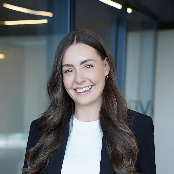 Frau mit langen braunen Haaren in Anzug steht mit verschränkten Armen vor einem Fenster.