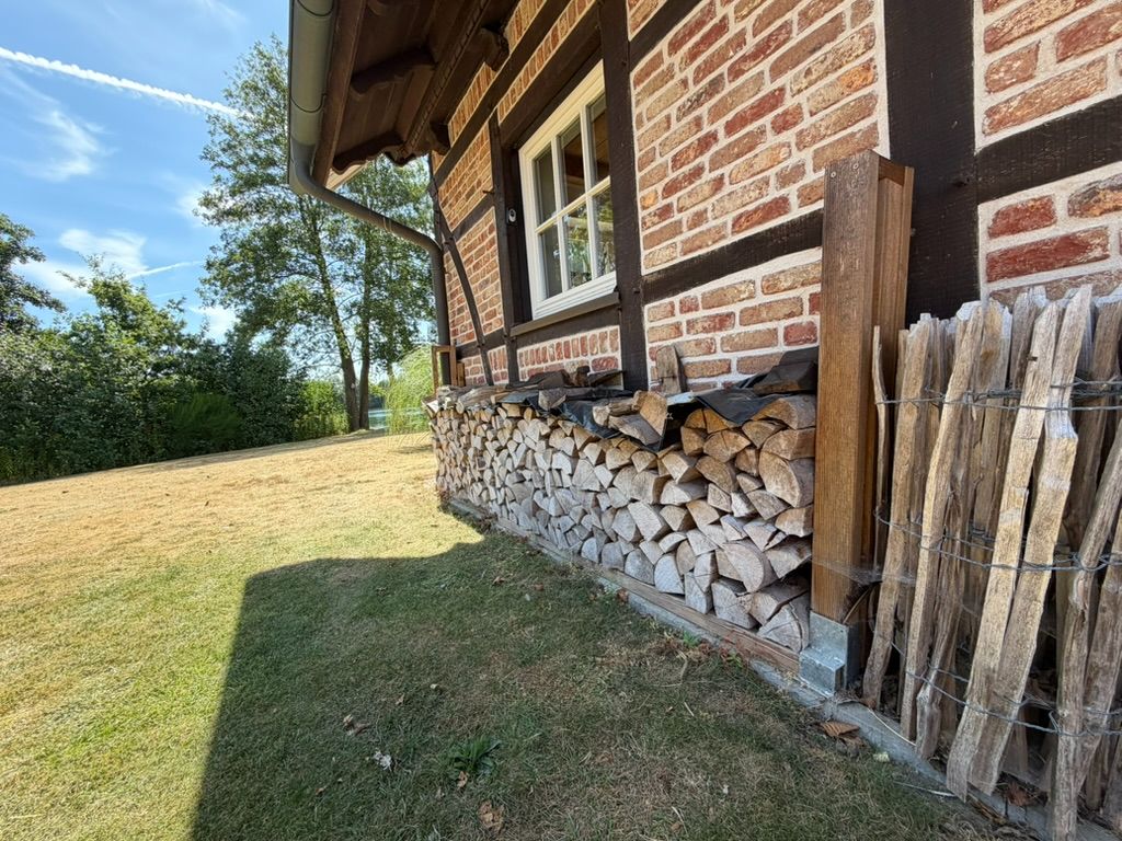 Gestapeltes Brennholz liegt an einer Ziegelhauswand mit Fenster im Garten.