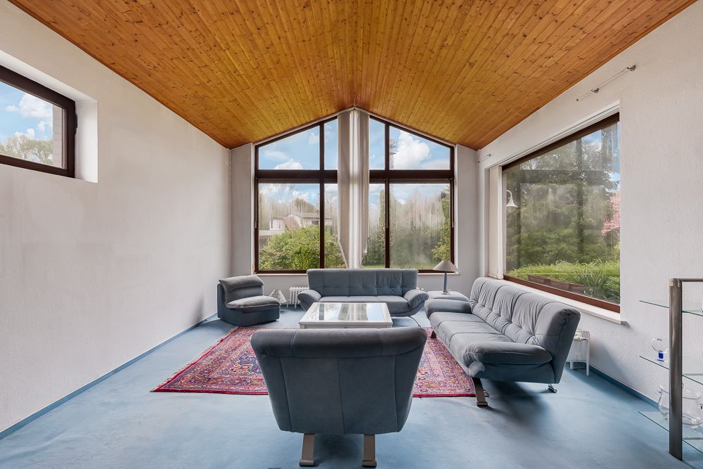 Wohnzimmer mit blauen Sofas, Glastisch, Holzbalkendecke und großen Fenstern zum Garten.