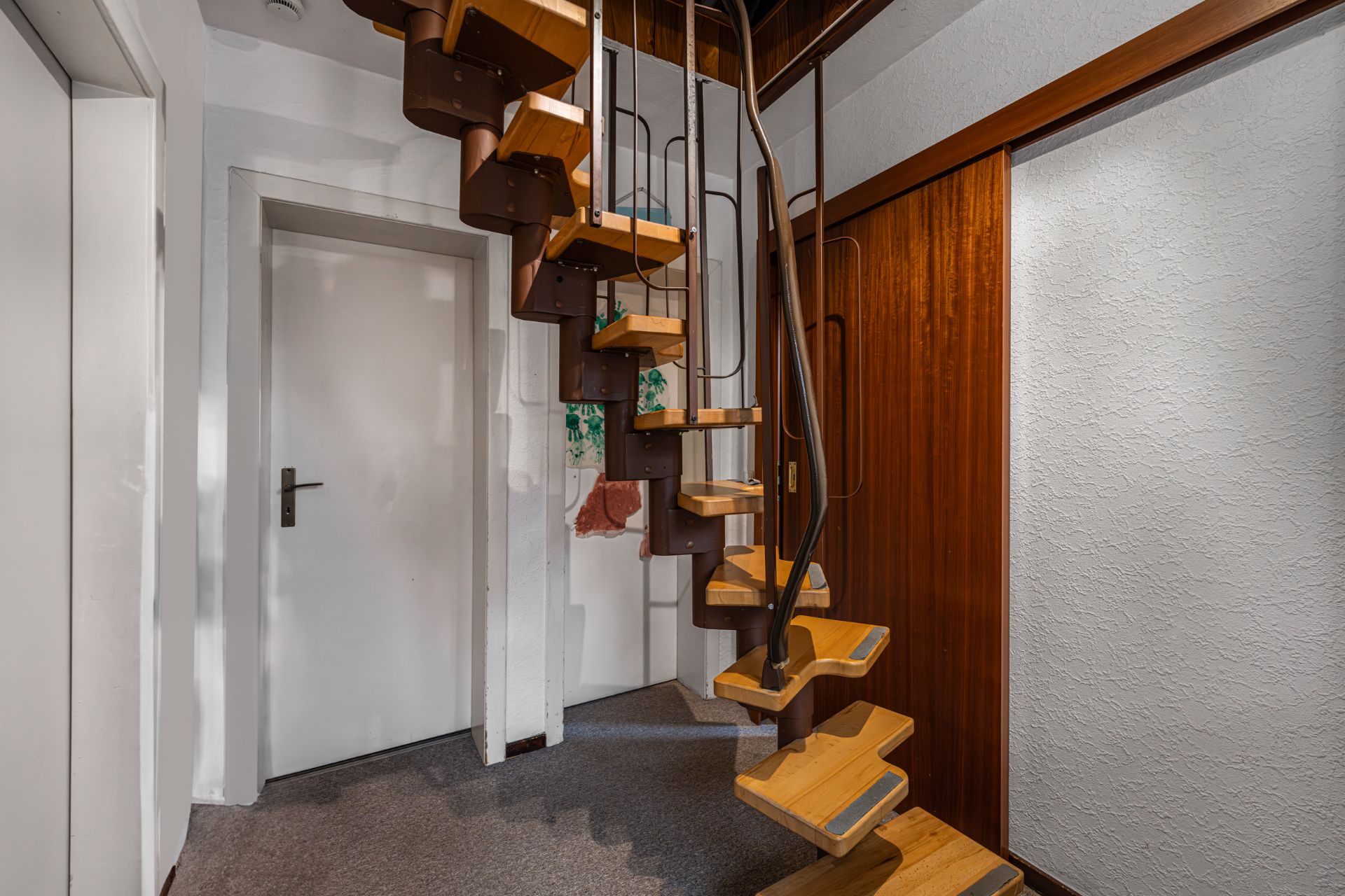 Wendeltreppe aus Holz und Metall in einem Flur mit weißen Wänden und Holztür.