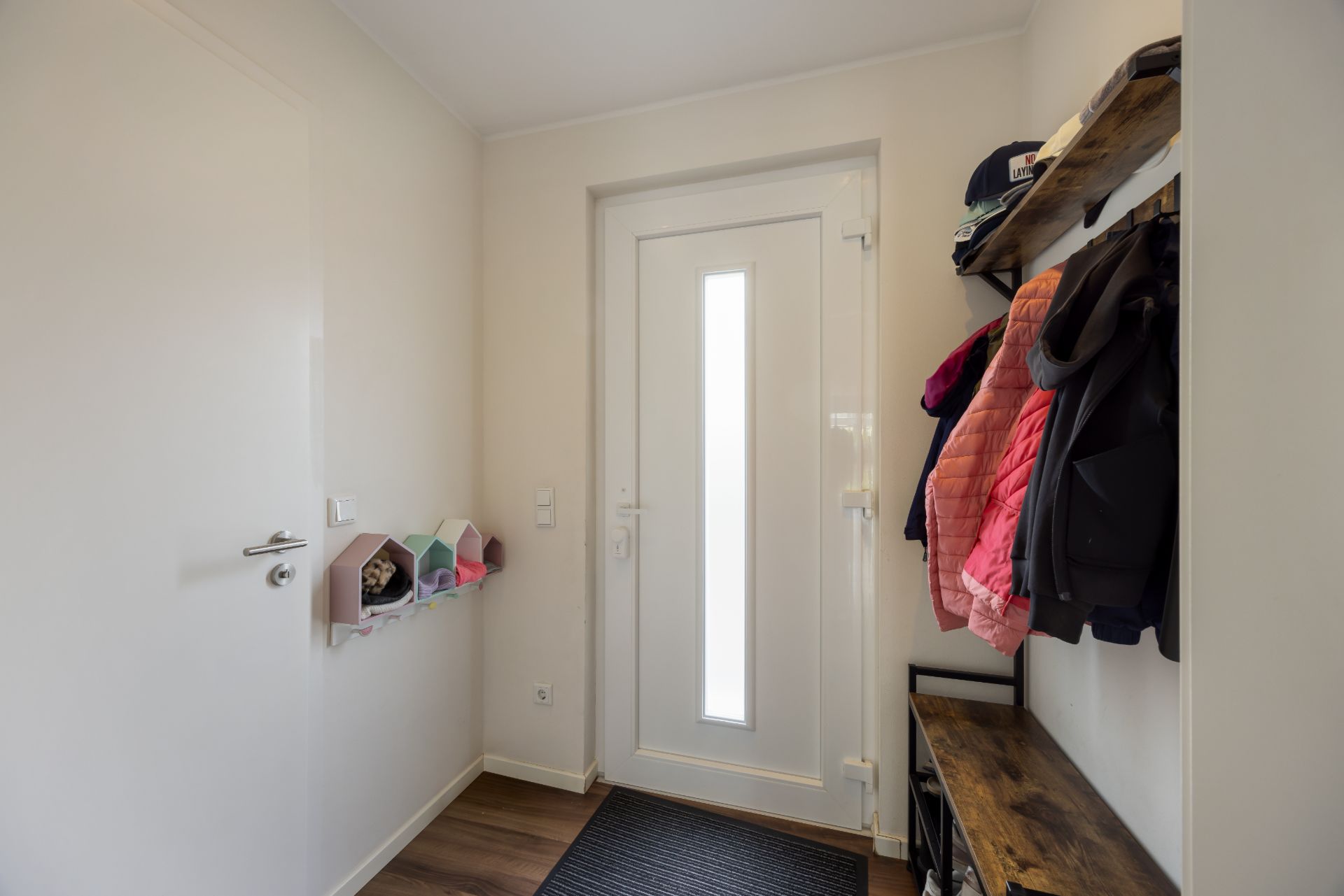 Flur mit weißer Haustür, Garderobe mit Jacken, Regal und kleiner Sitzbank.