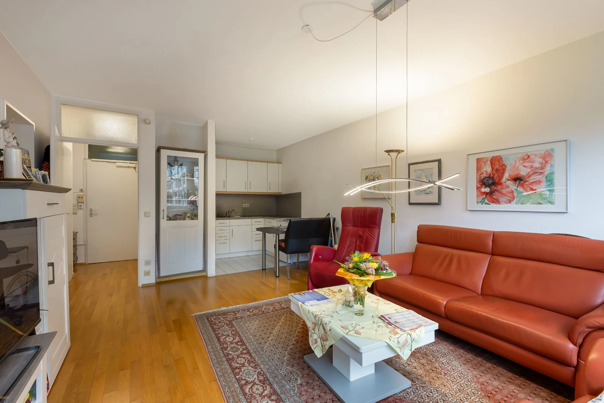 Helles Wohnzimmer mit rotem Sofa, Sessel, Teppich und offener Küche im Hintergrund.