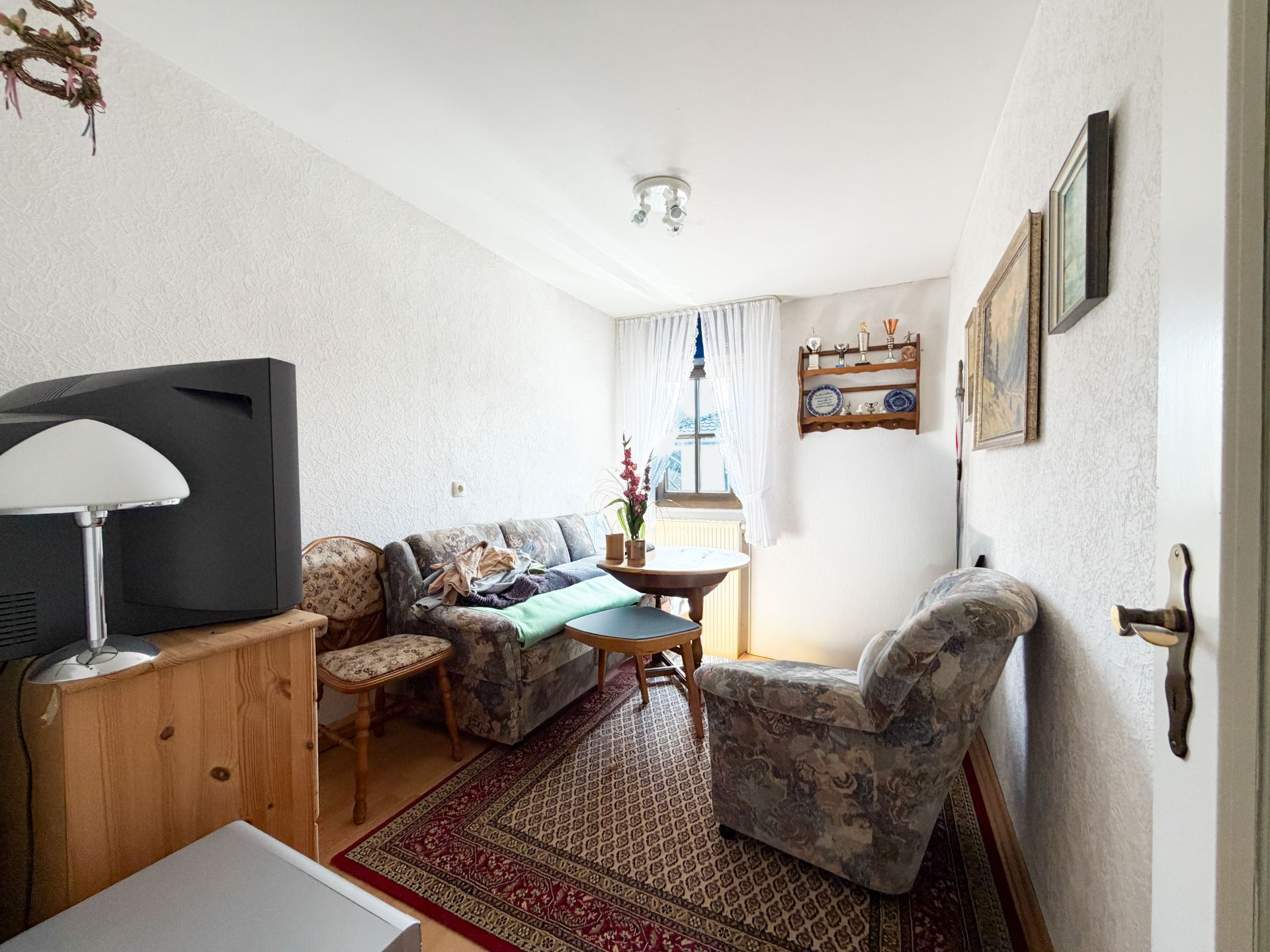Kleines Wohnzimmer mit gemustertem Teppich, Sofa, Sessel, altem Fernseher und Fenster mit Vorhängen.