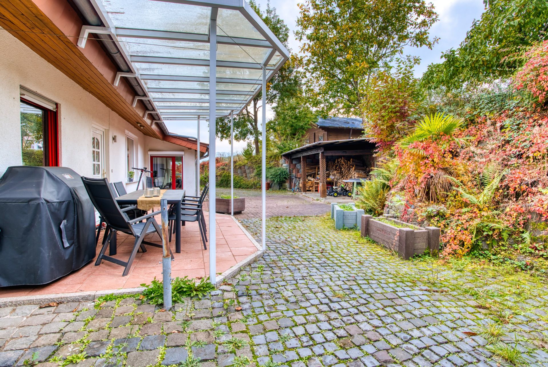 Überdachte Terrasse mit Tisch, Stühlen und Grill, daneben Hof mit Pflanzen und Schuppen.