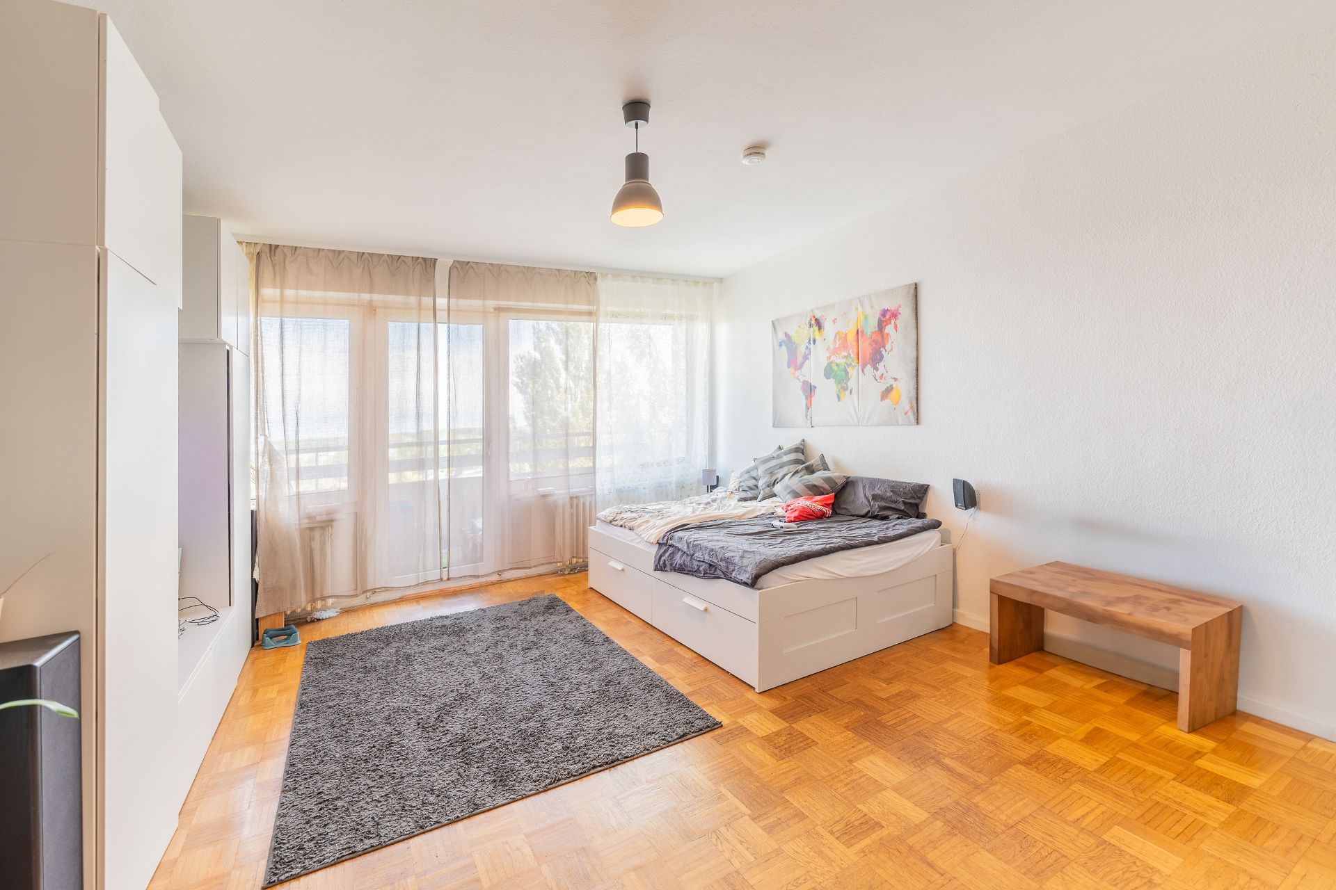 Helles Schlafzimmer mit Doppelbett, Teppich, Bank und großem Fenster mit Vorhängen.