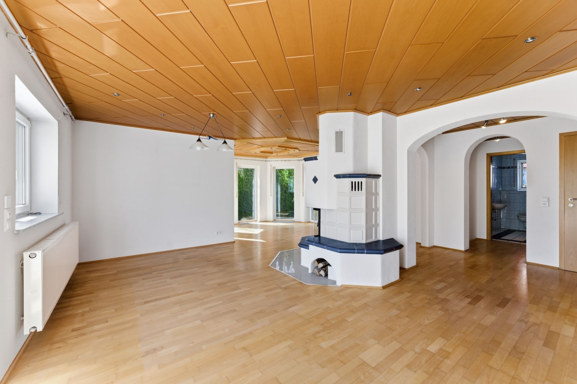 Helles Wohnzimmer mit Holzdecke, Kaminofen und Zugang zu weiteren Räumen, Holzboden und großen Fenstern.