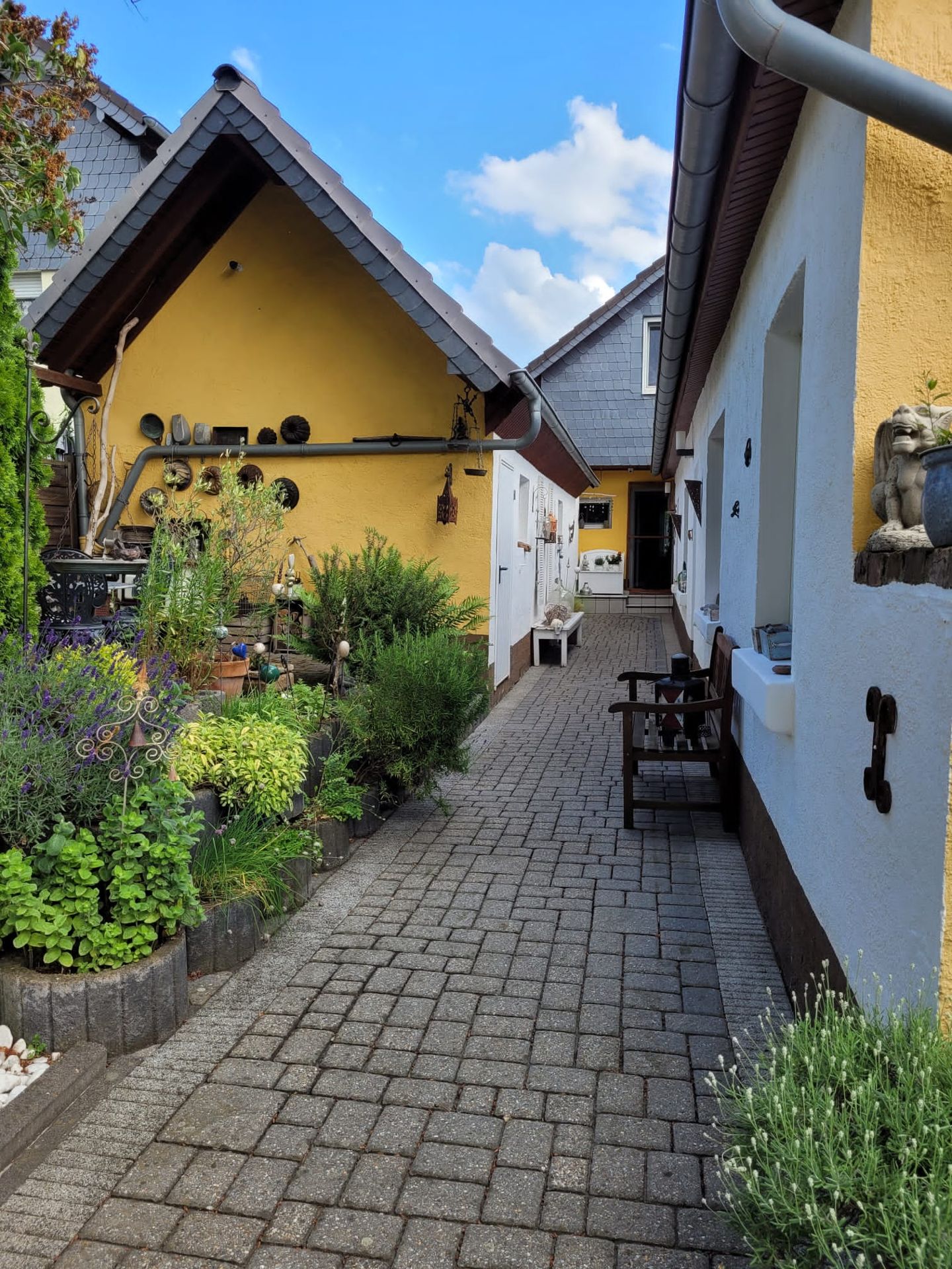 Gepflasterter Weg zwischen gelbem Haus und Garten mit Pflanzen, Bänken und Dekoration.