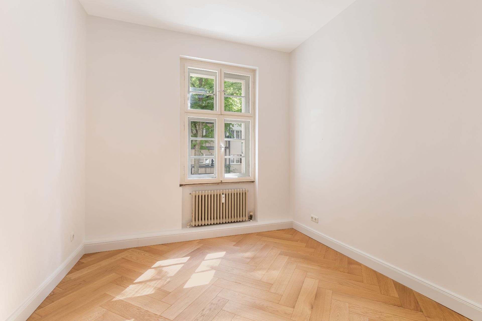 Heller, leerer Raum mit Parkettboden, weiß gestrichenen Wänden und einem Fenster mit Heizkörper.