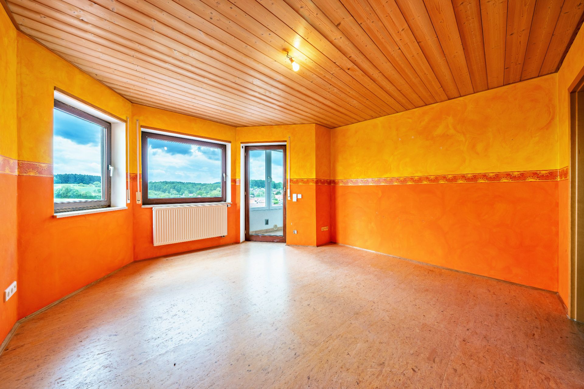 Leeres Zimmer mit orange-gelben Wänden, Holzboden und Decke, mehrere Fenster und Tür zum Balkon.