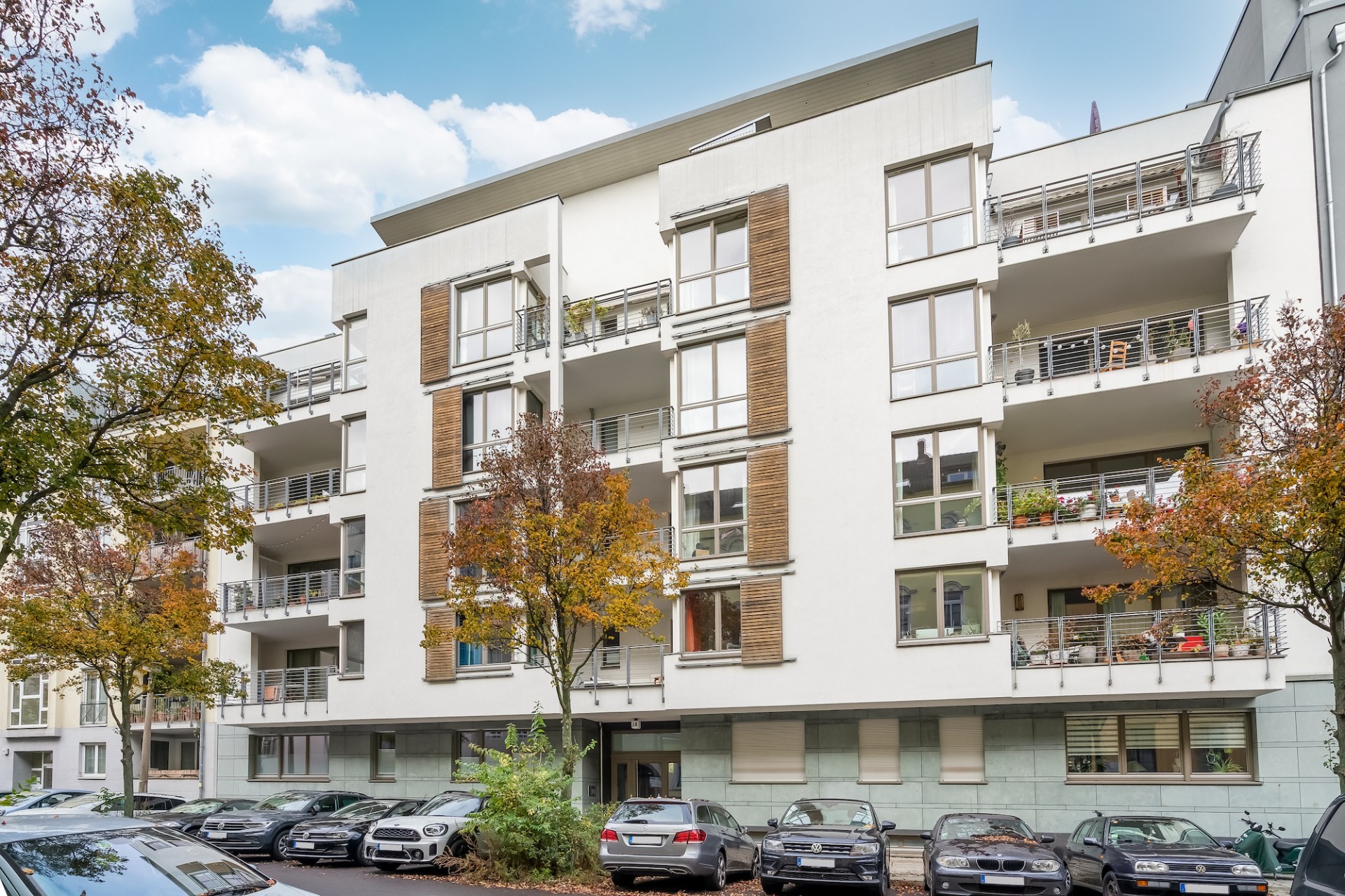Modernes Wohnhaus mit Balkonen und geparkten Autos davor, umgeben von Bäumen im Herbst.