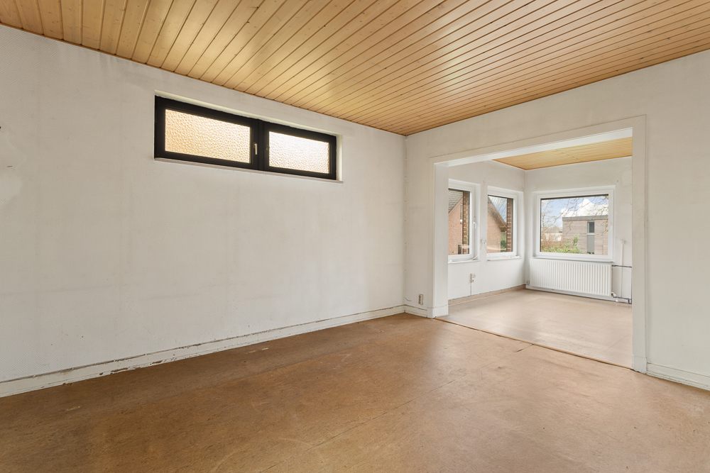 Leerer Raum mit Holzdecke, braunem Boden und zwei Fenstern, angrenzend an einen weiteren Raum mit großen Fenstern.