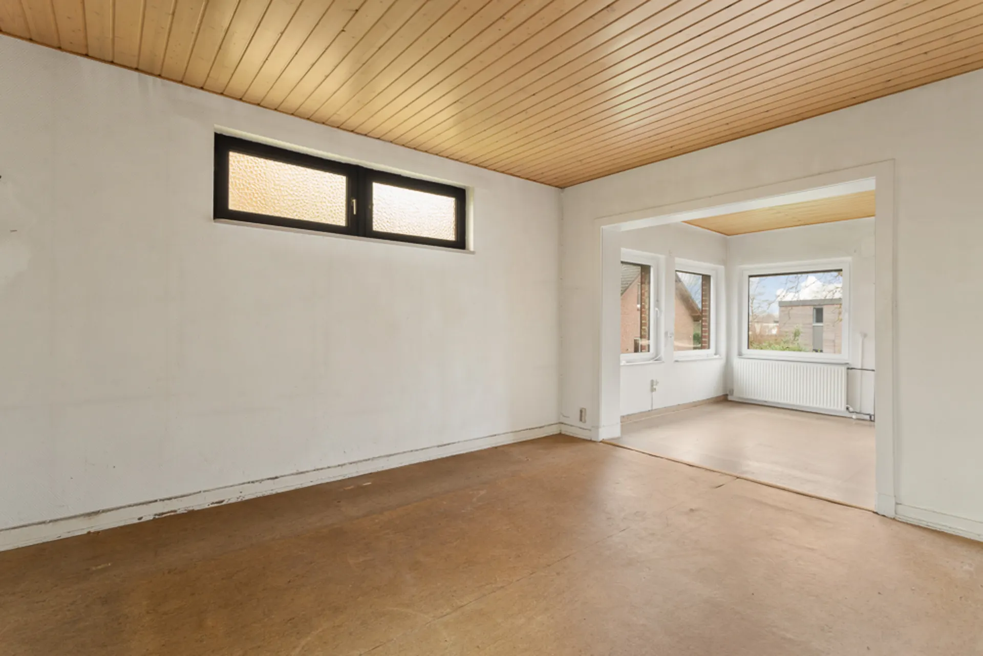 Leerer Raum mit Holzdecke, braunem Boden und zwei Fenstern, angrenzend an einen weiteren Raum mit großen Fenstern.