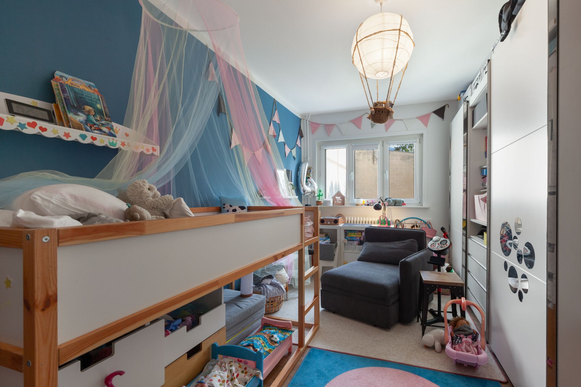 Kinderzimmer mit Hochbett, Spielsachen, Sessel und Fenster, bunte Dekorationen an der Wand.