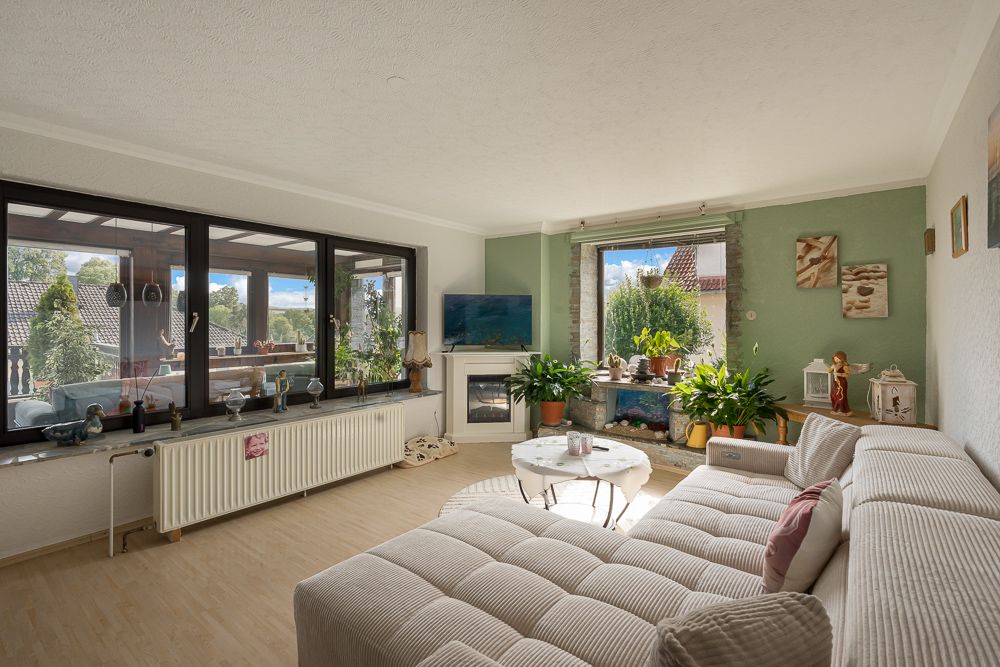 Helles Wohnzimmer mit großer Eckcouch, Couchtisch, vielen Pflanzen und großen Fenstern.