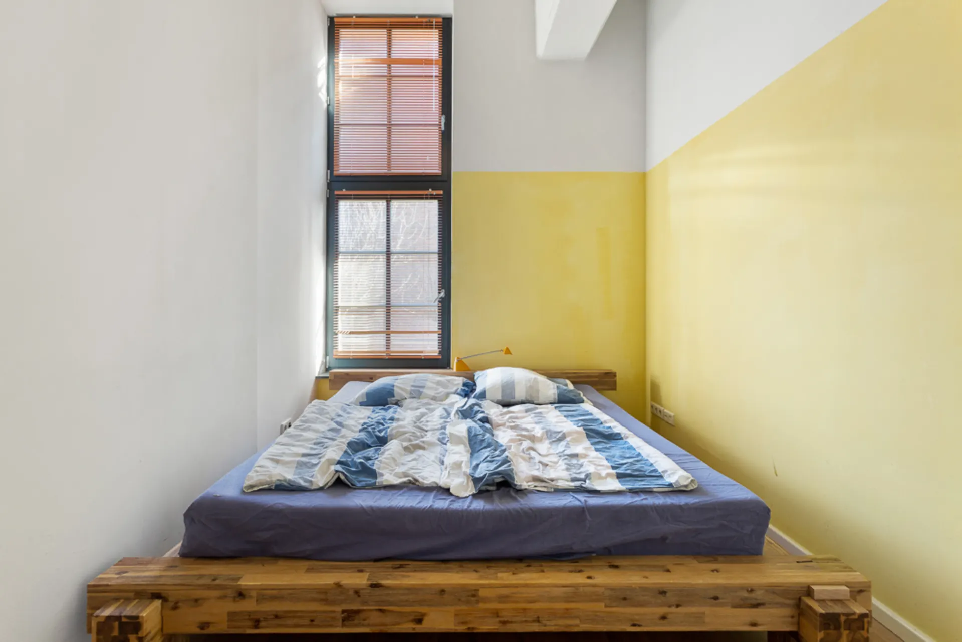 Schlafzimmer mit Holzbett, bunter Bettwäsche und hohem Fenster mit Jalousien.