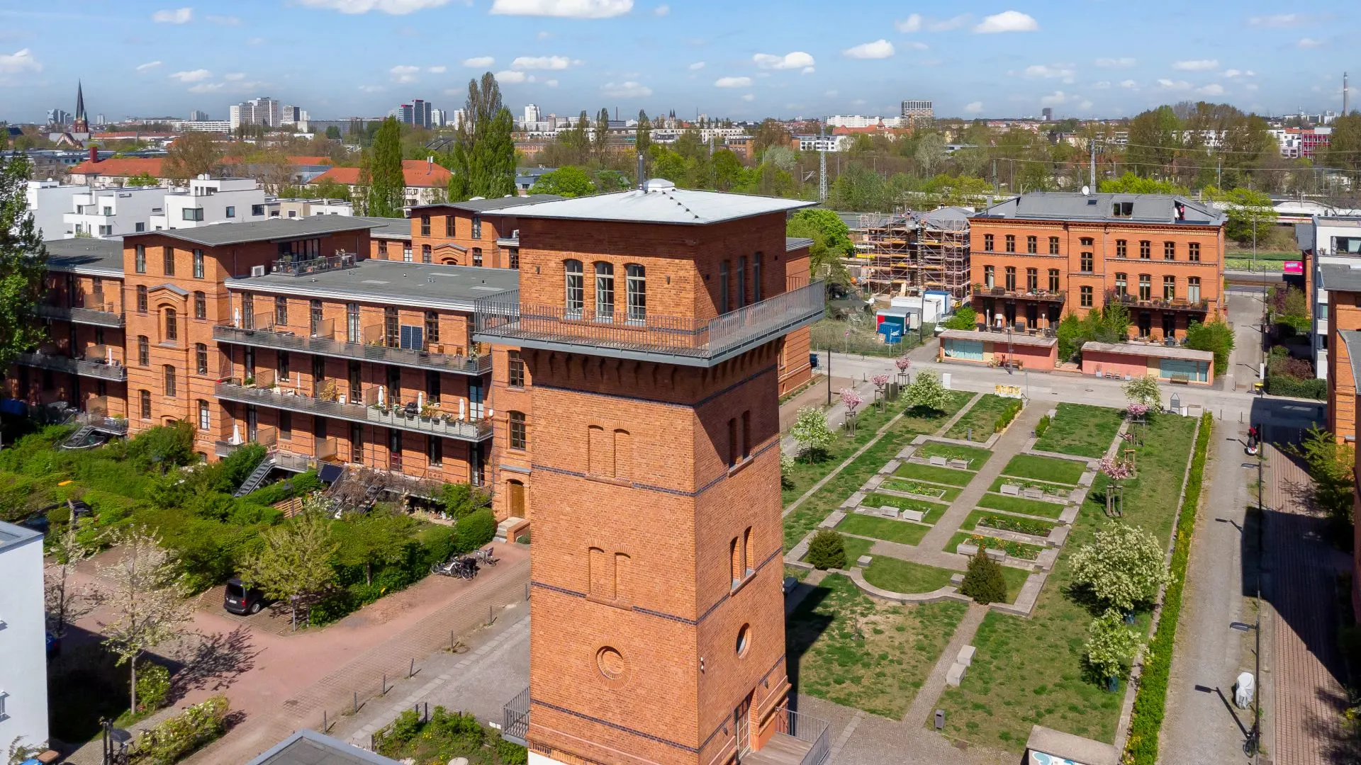 Backsteingebäude mit Innenhof und Gartenanlage, im Vordergrund ein Turm, umgeben von Wegen und Bäumen.
