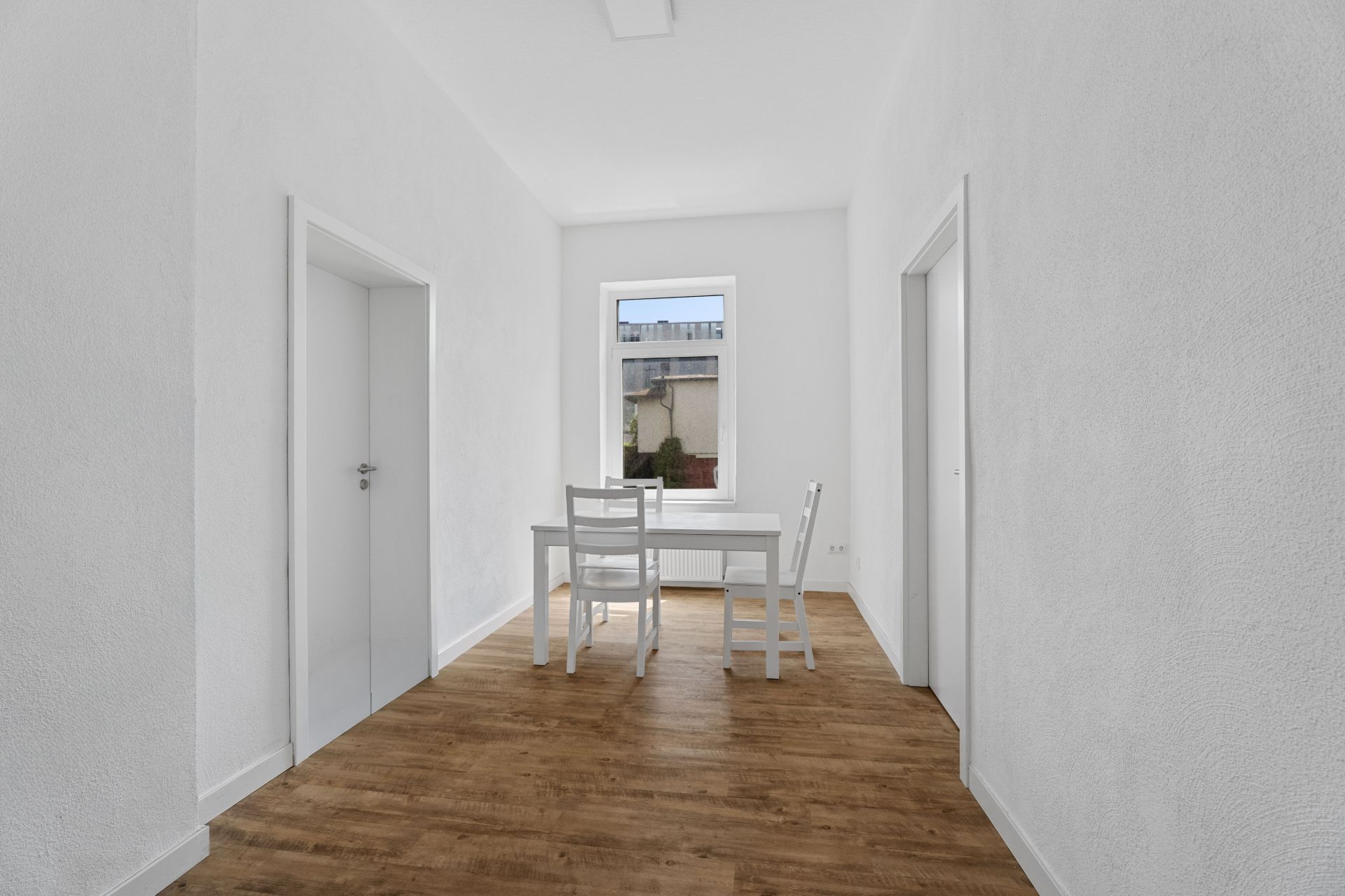 Heller Raum mit Holzboden, einem Tisch und drei weißen Stühlen vor einem Fenster.