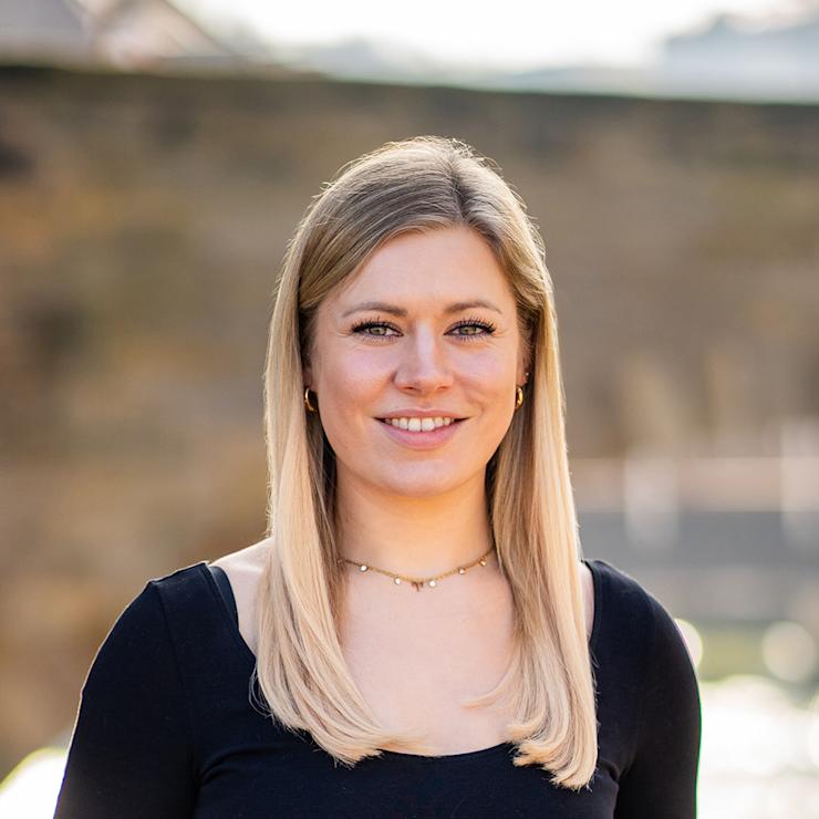 Eine Frau mit langen blonden Haaren steht draußen vor einer unscharfen Hintergrundmauer.