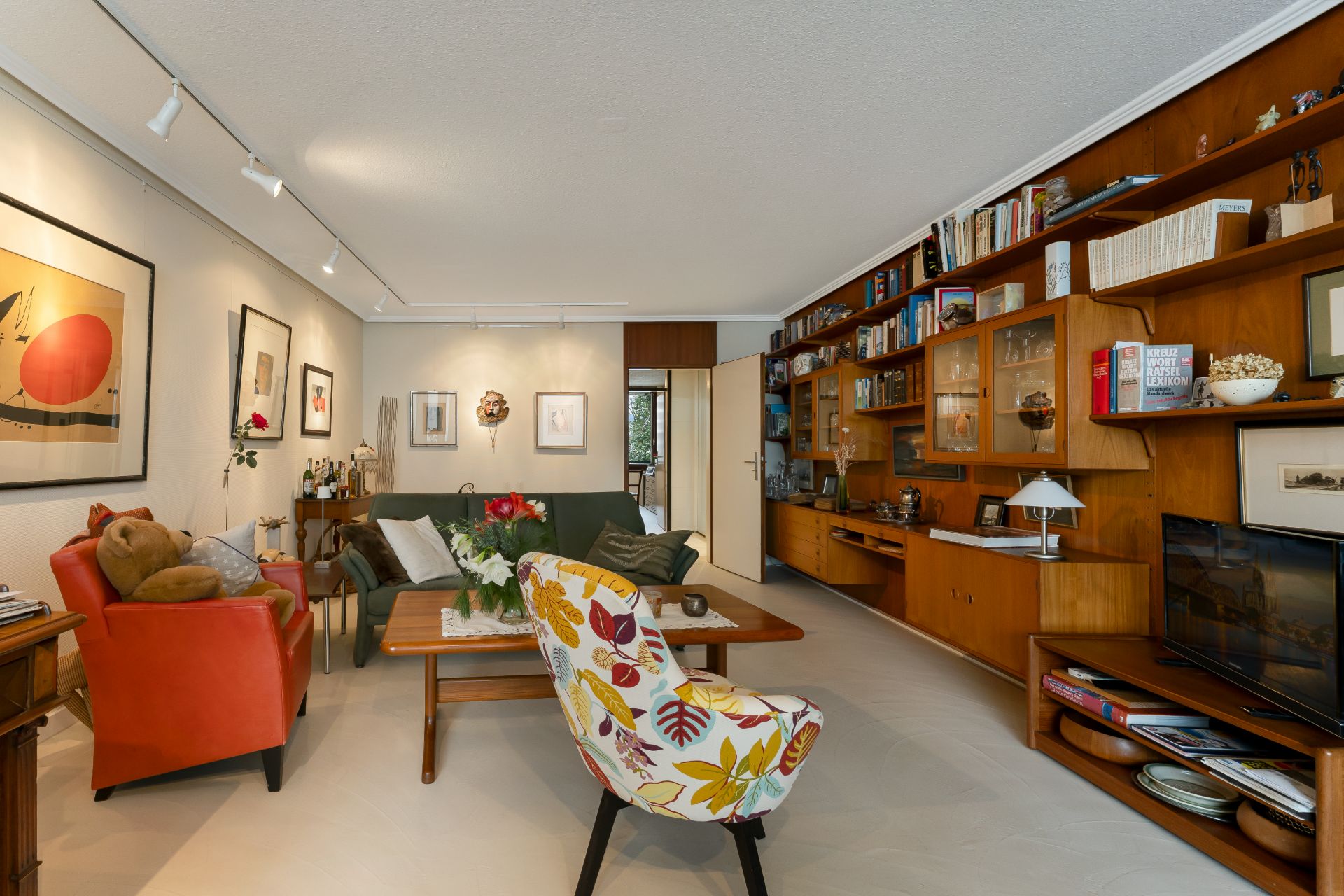 Wohnzimmer mit buntem Sessel, rotem Sofa, Holzmöbeln und Bücherregal an der Wand.