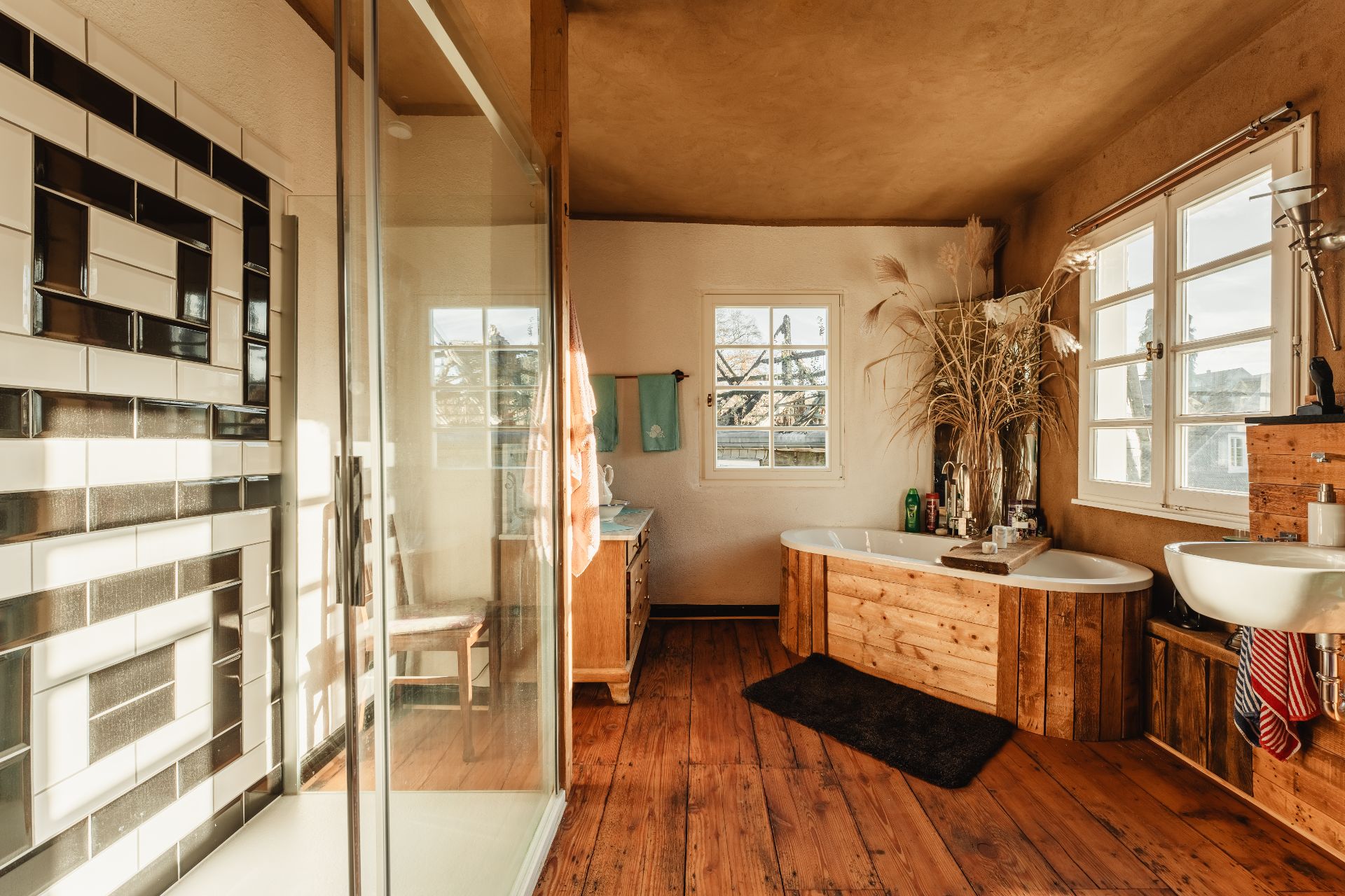Helles Badezimmer mit Holzfußboden, Badewanne, Dusche, Waschbecken und großen Fenstern.