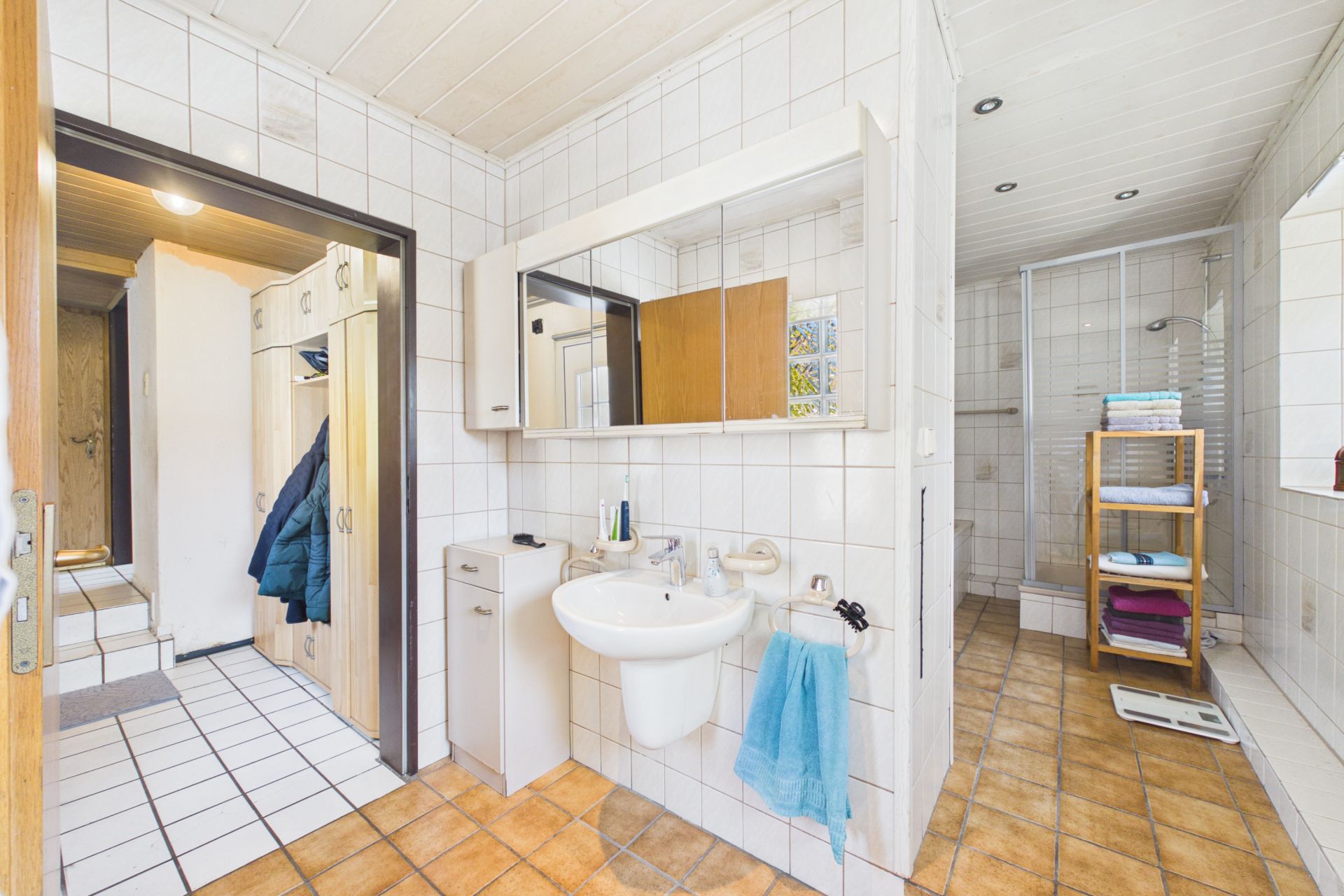 Helles Badezimmer mit Waschbecken, Spiegel, Dusche und Regal mit Handtüchern.
