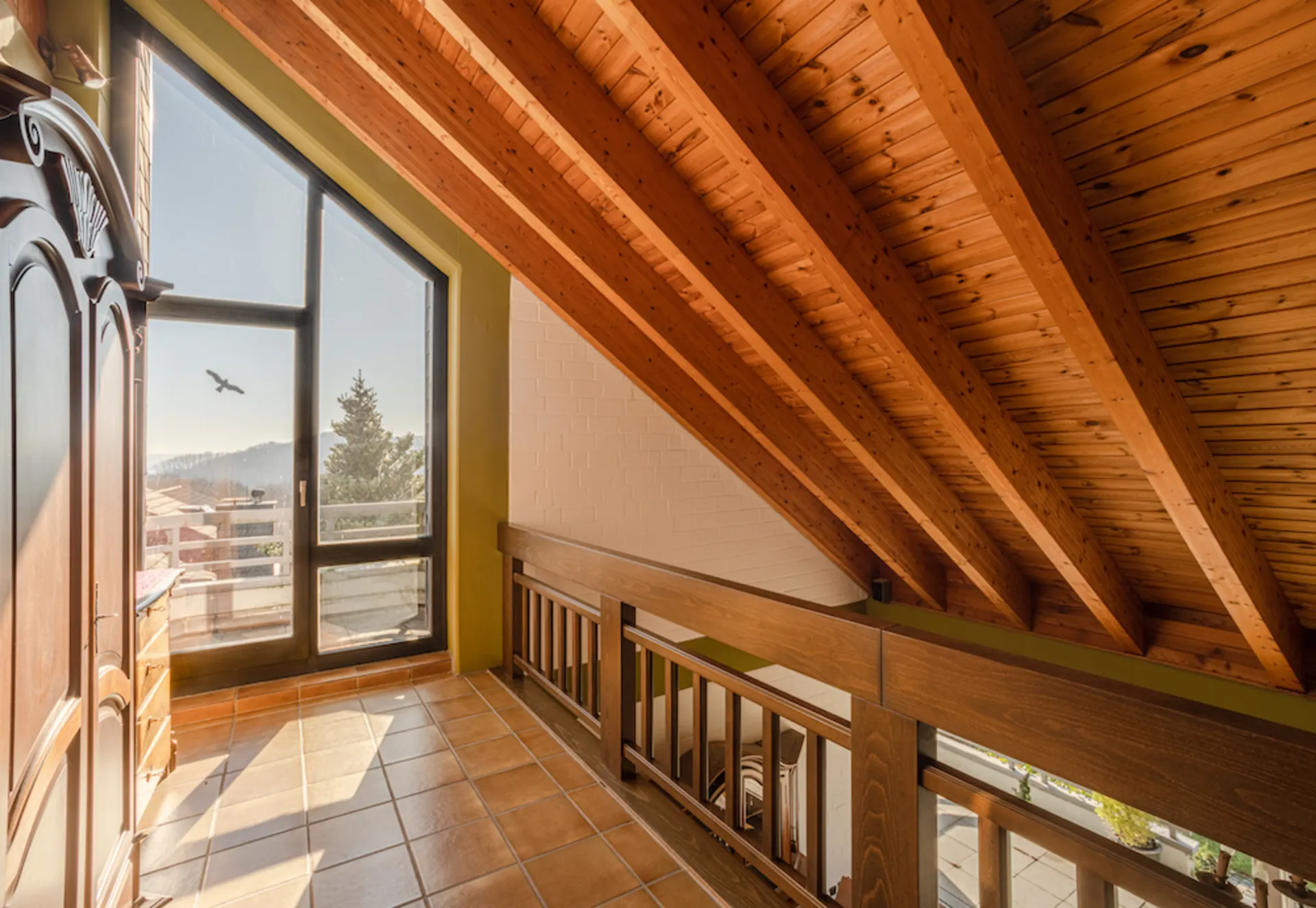 Helles Dachgeschoss mit Holzdecke, Fliesenboden und großem Fenster mit Aussicht nach draußen.