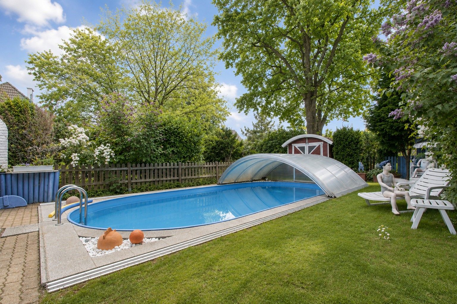 Garten mit Swimmingpool, teilweise überdacht, Liegestühlen und Bäumen im Hintergrund.