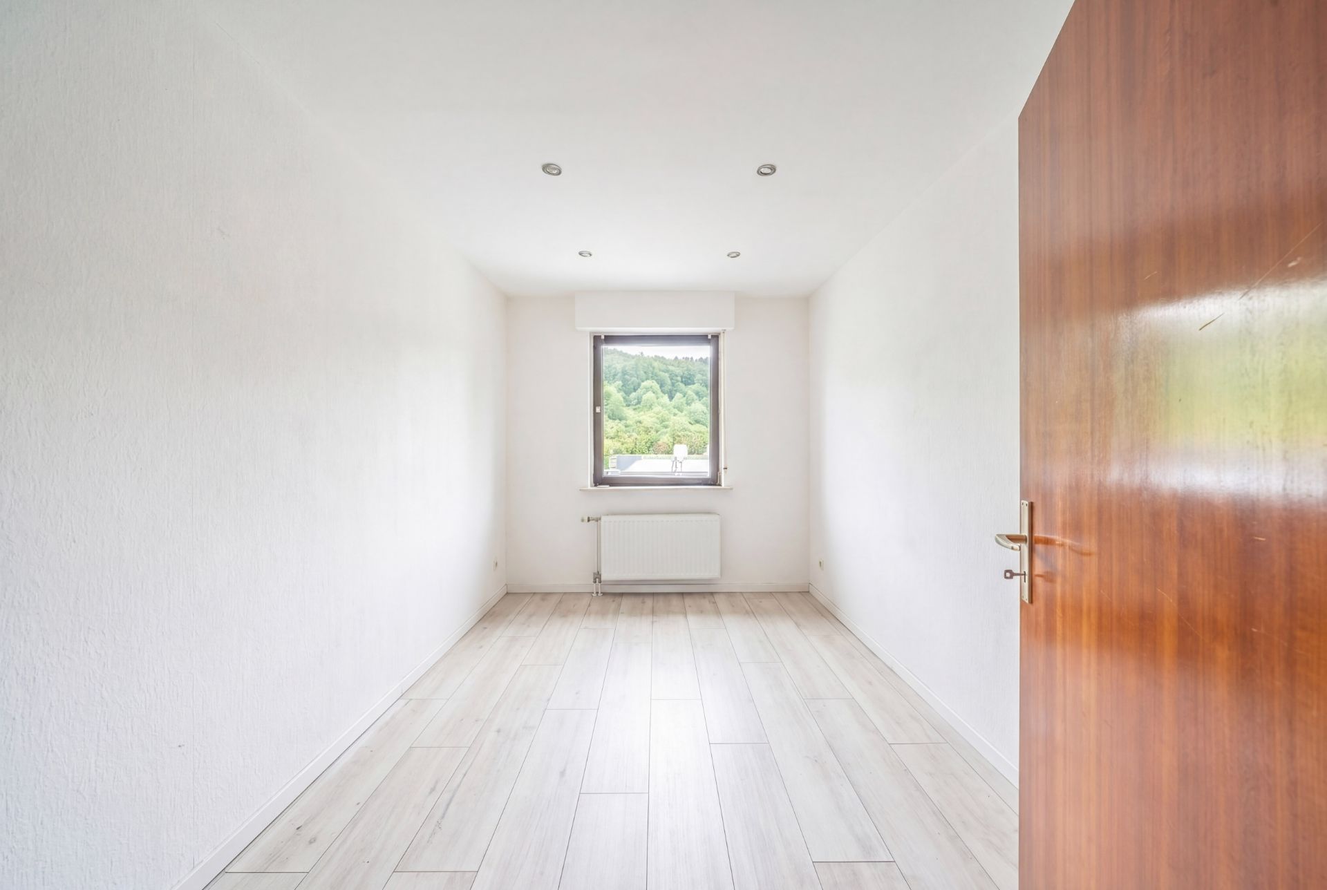 Leerer, heller Raum mit Holzboden, weißem Fenster und Blick ins Grüne.