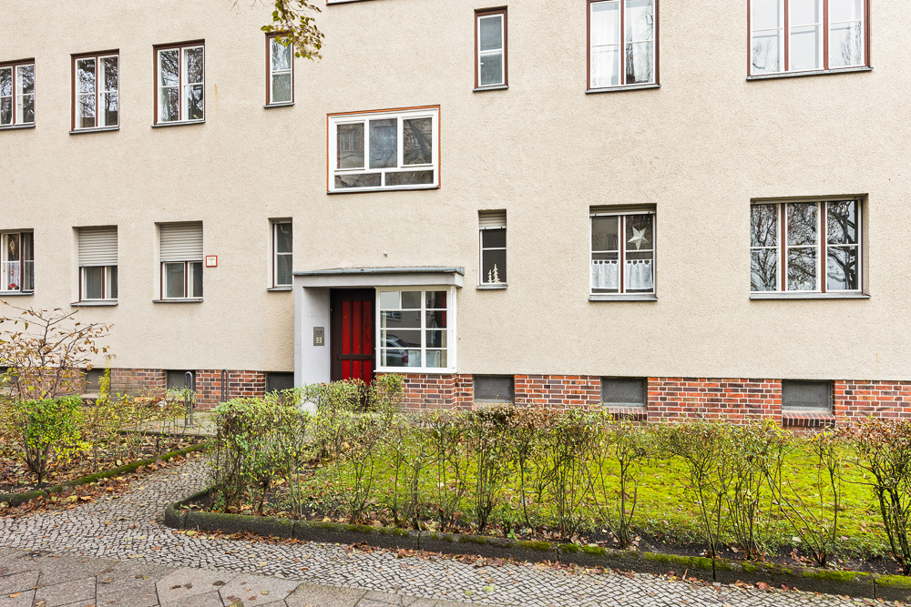 Eingang eines Mehrfamilienhauses mit roter Tür und mehreren Fenstern, davor grüne Hecke.