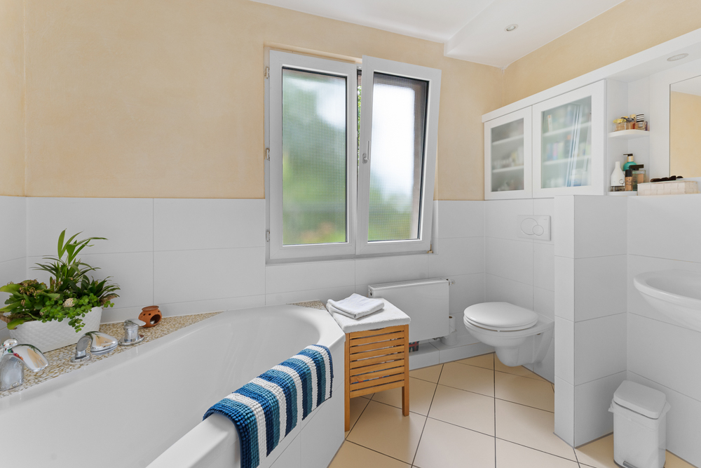 Helles Badezimmer mit Badewanne, Pflanze, Fenster, WC und Handtuch auf einem Hocker.