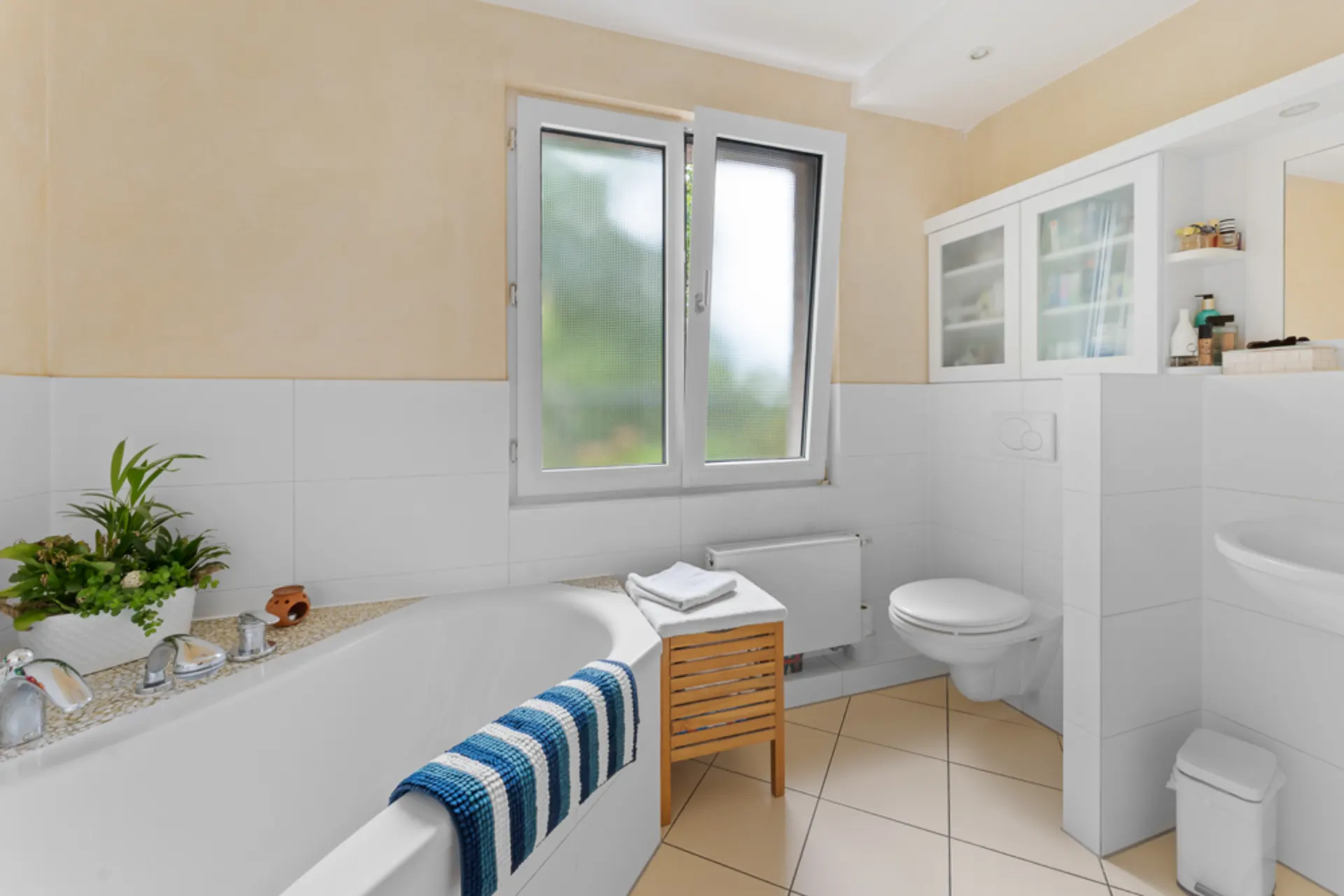 Helles Badezimmer mit Badewanne, Pflanze, Fenster, WC und Handtuch auf einem Hocker.