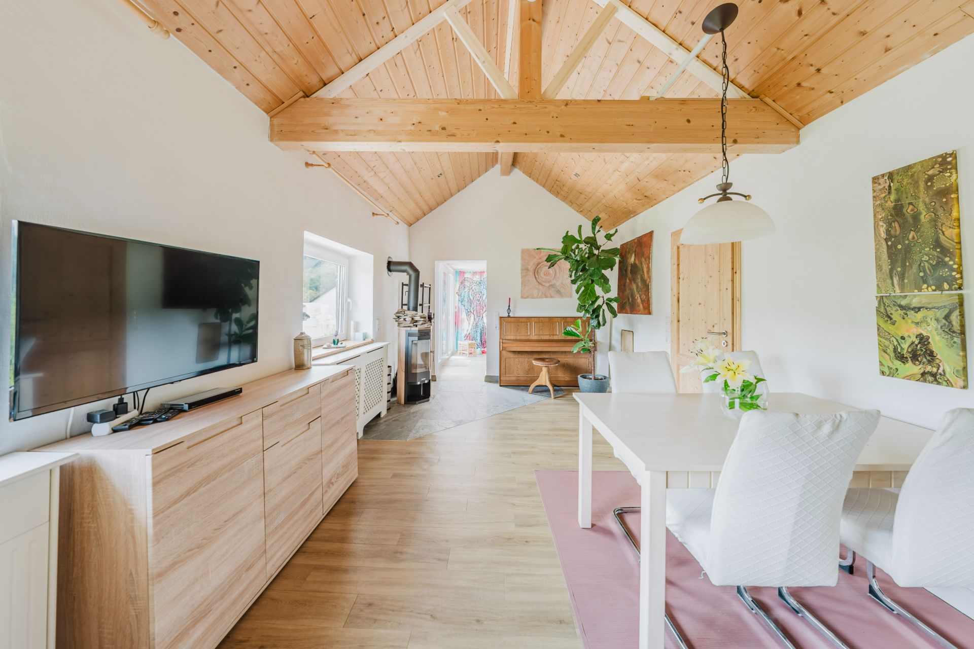 Helles Wohnzimmer mit Holzbalkendecke, Esstisch, Fernseher und Pflanzen.
