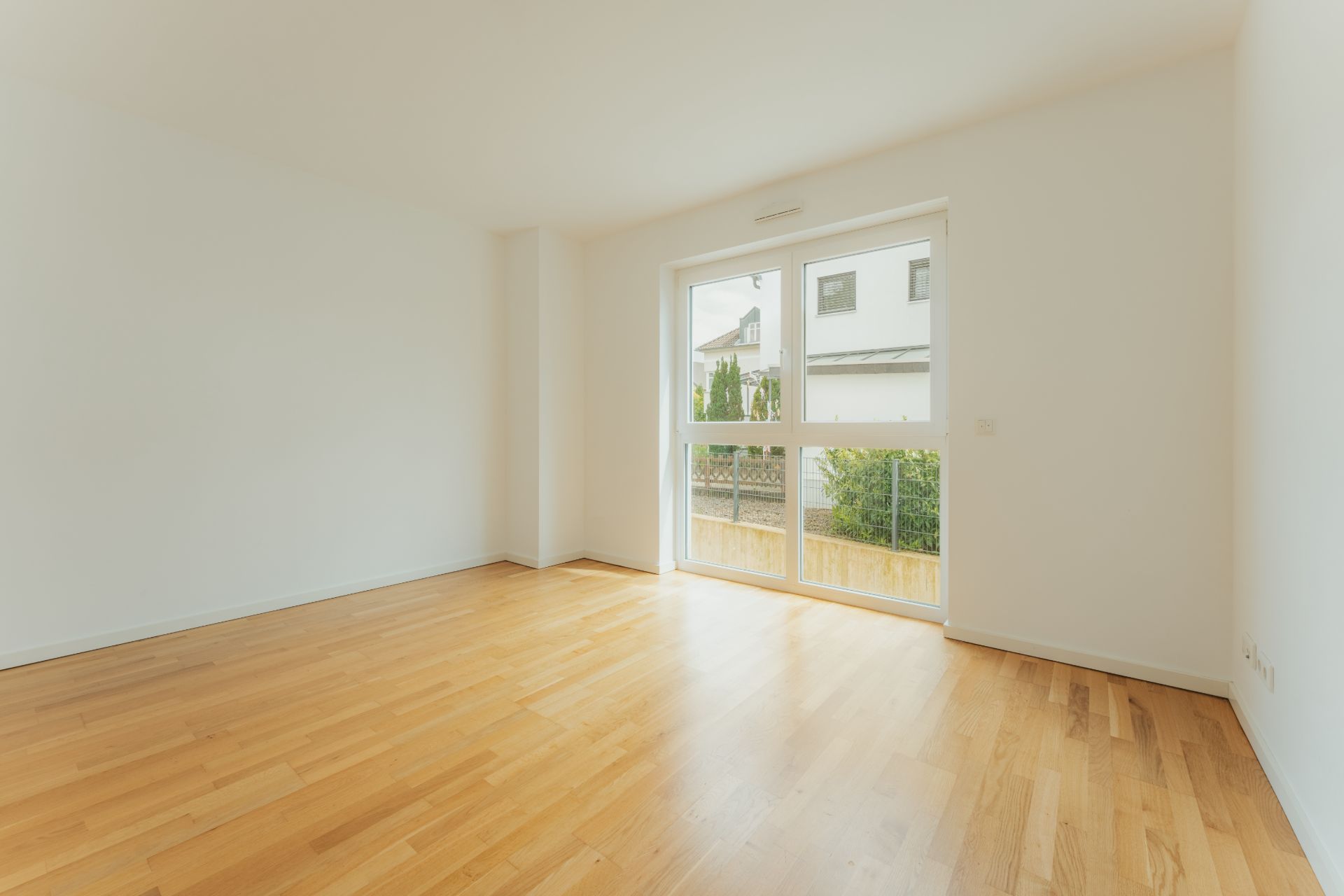 Helles, leeres Zimmer mit Parkettboden und großem Fenster, Blick nach draußen auf Garten und Nachbarhaus.