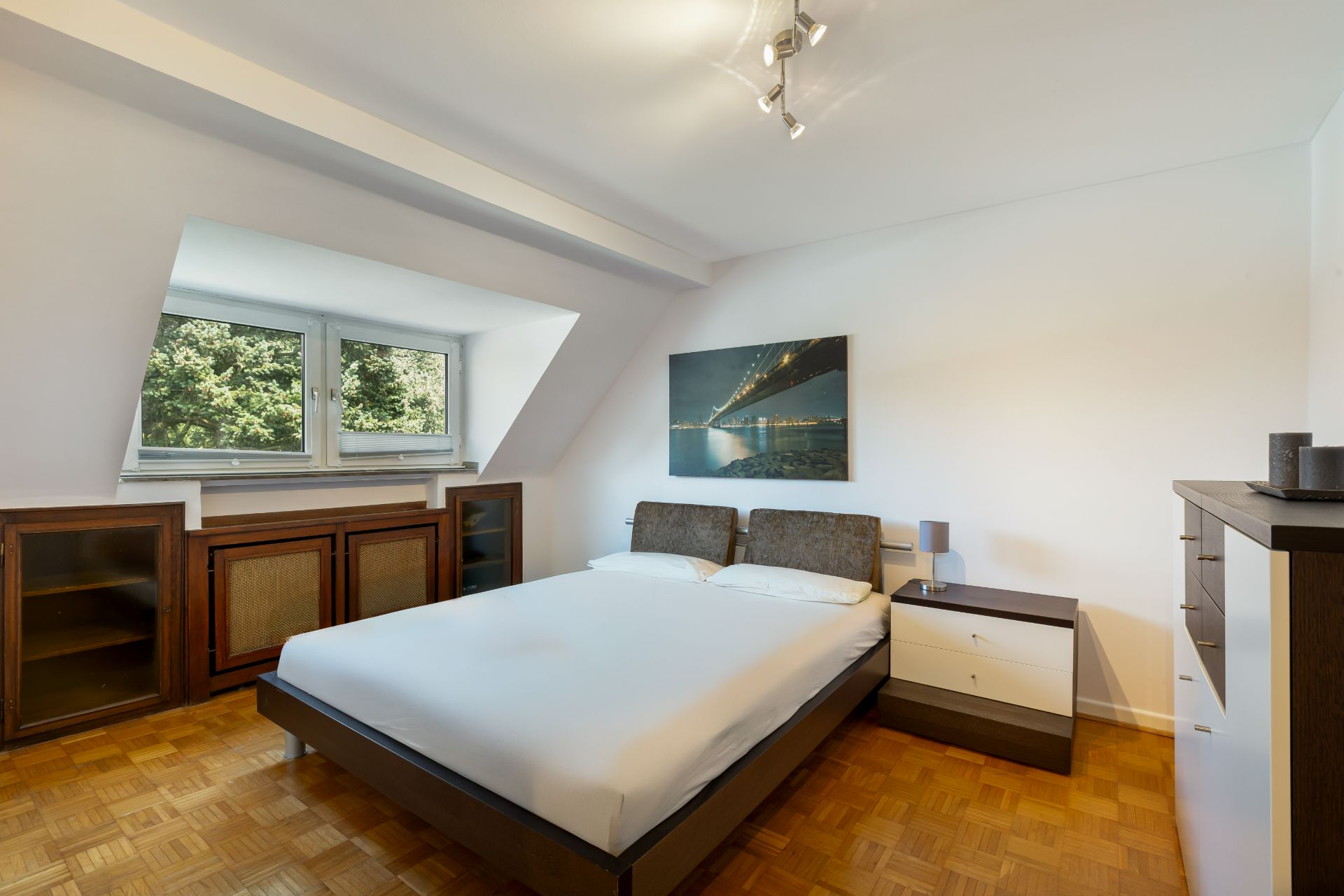 Helles Schlafzimmer mit Doppelbett, zwei Nachttischen, Kommode und Fenstern mit Blick ins Grüne.